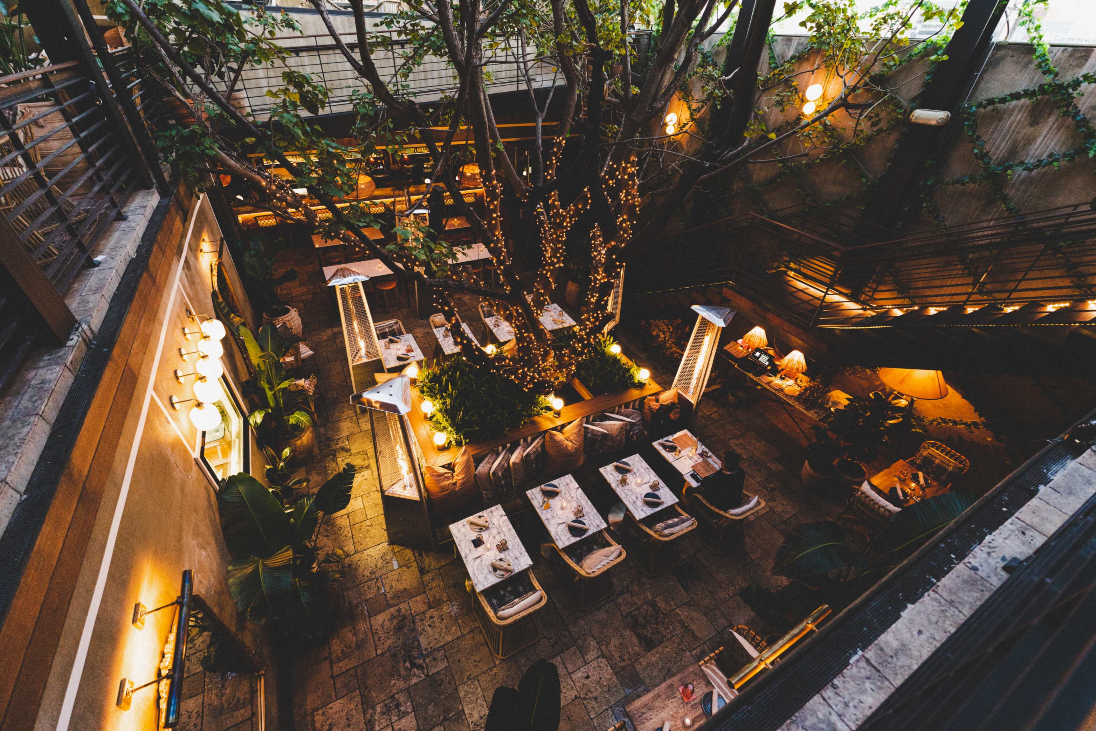 A sunken restaurant features outdoor seating surrounded by greenery and string lights illuminating the space.