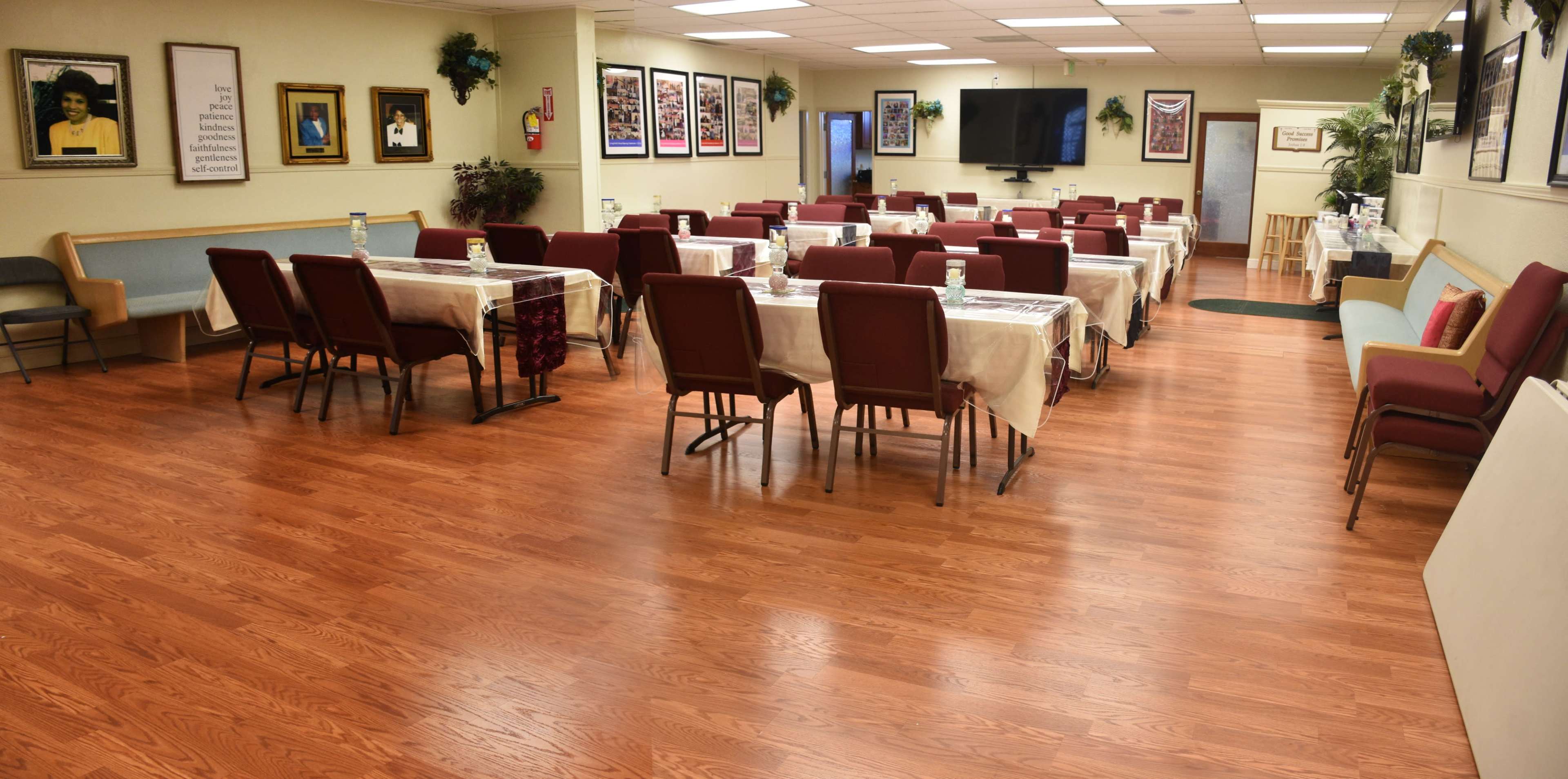 A spacious dining area features rows of empty tables covered with white tablecloths and surrounded by red chairs, with framed photographs on the walls.