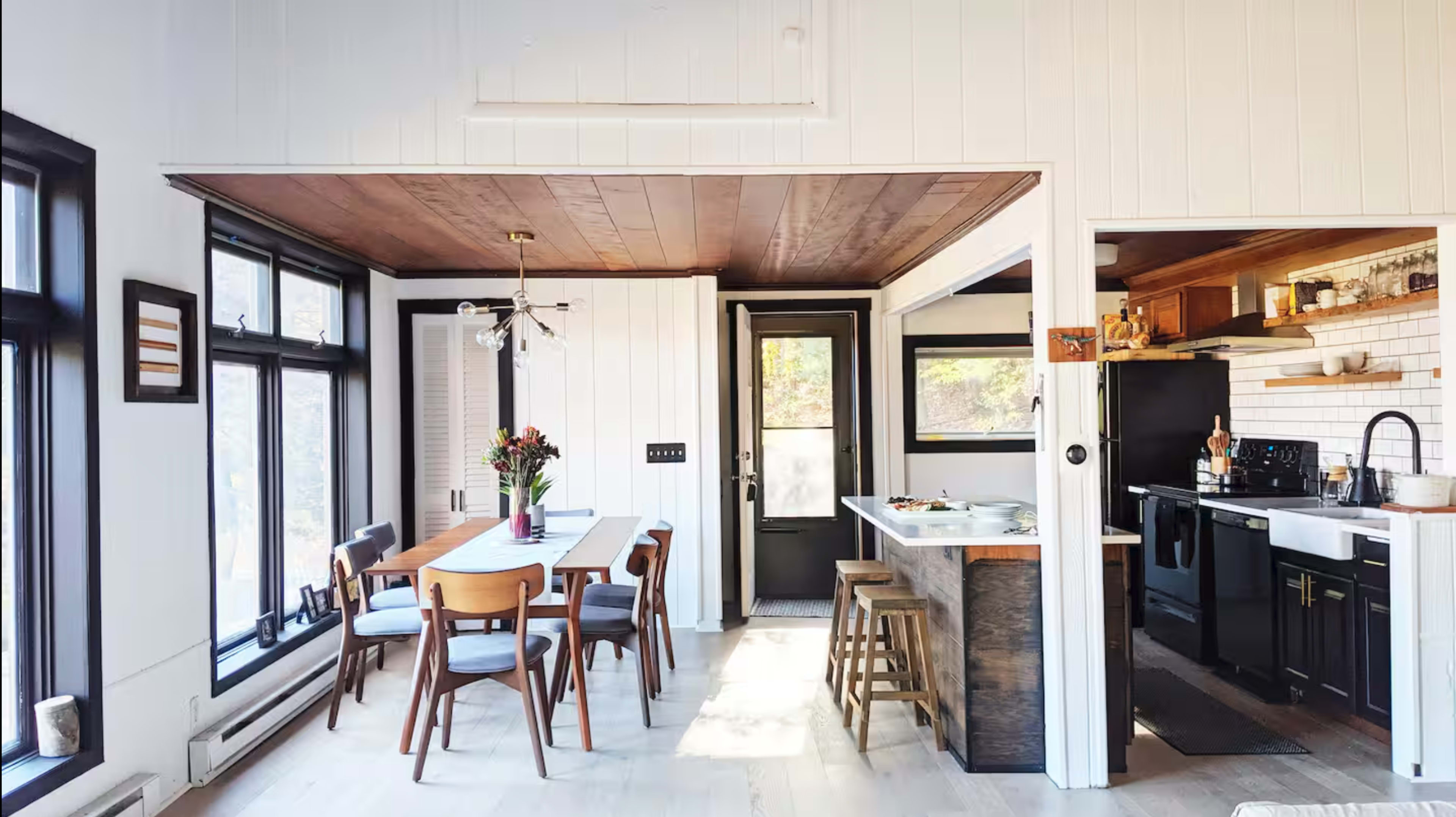 The image shows a modern dining area with a wooden table surrounded by chairs, leading into a kitchen with black appliances and wood accents.