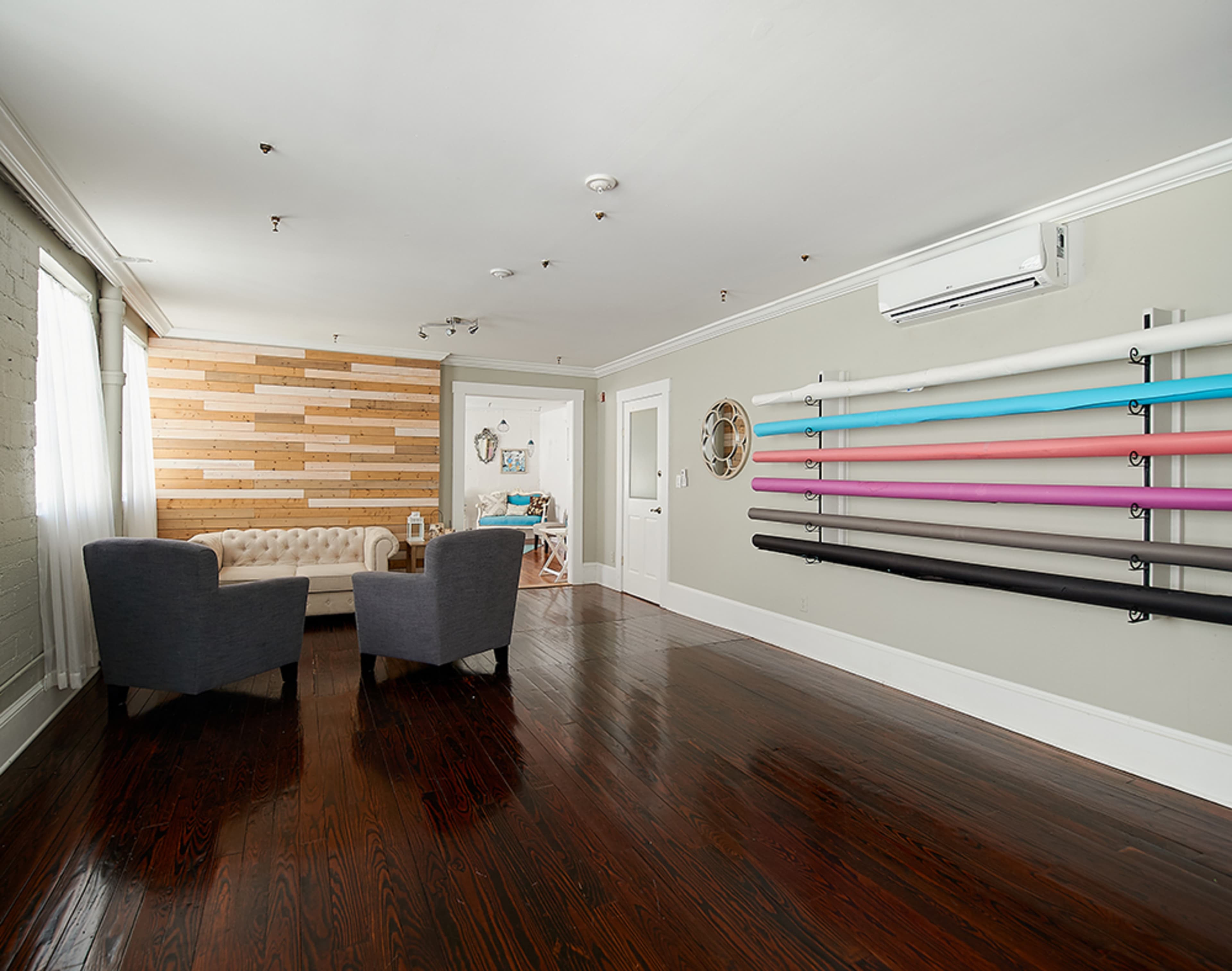 The image shows a spacious room with a wooden accent wall, two gray armchairs, and a wall-mounted display of colorful exercise bands.