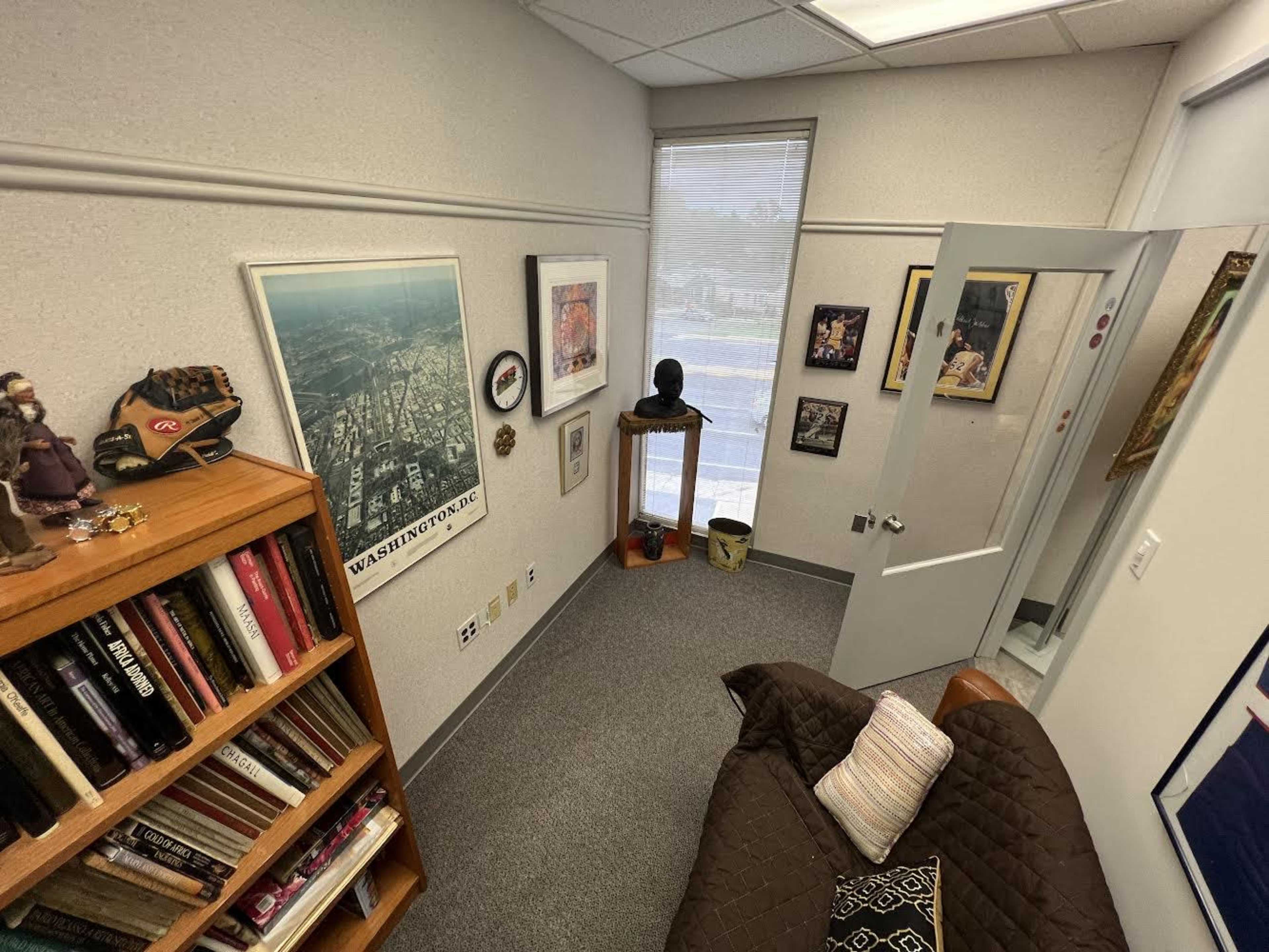 A small office space with a bookshelf filled with books, a couch, and various framed artworks on the walls.