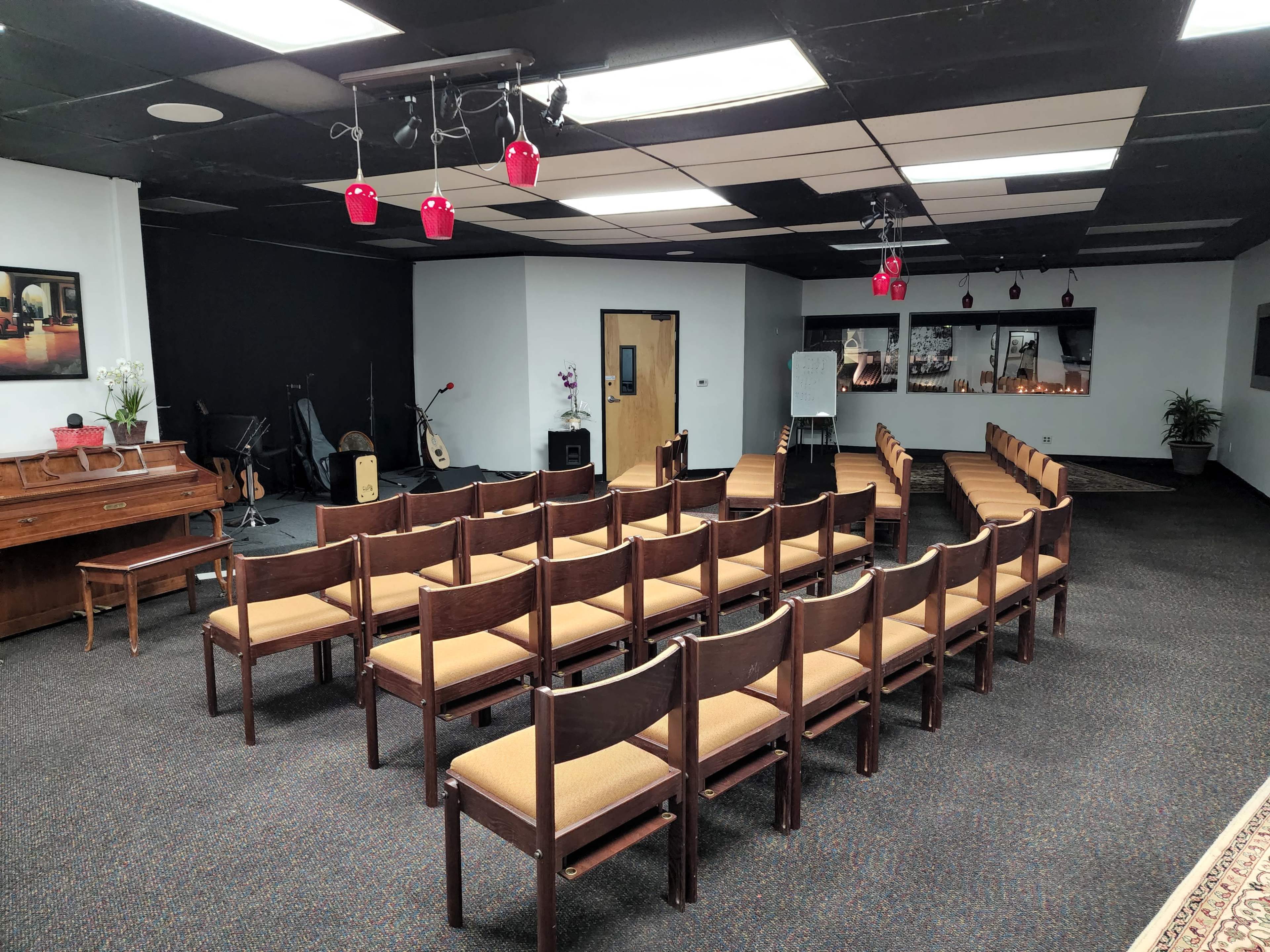 A room arranged with rows of wooden chairs facing a wall with a piano and musical instruments visible in the background.