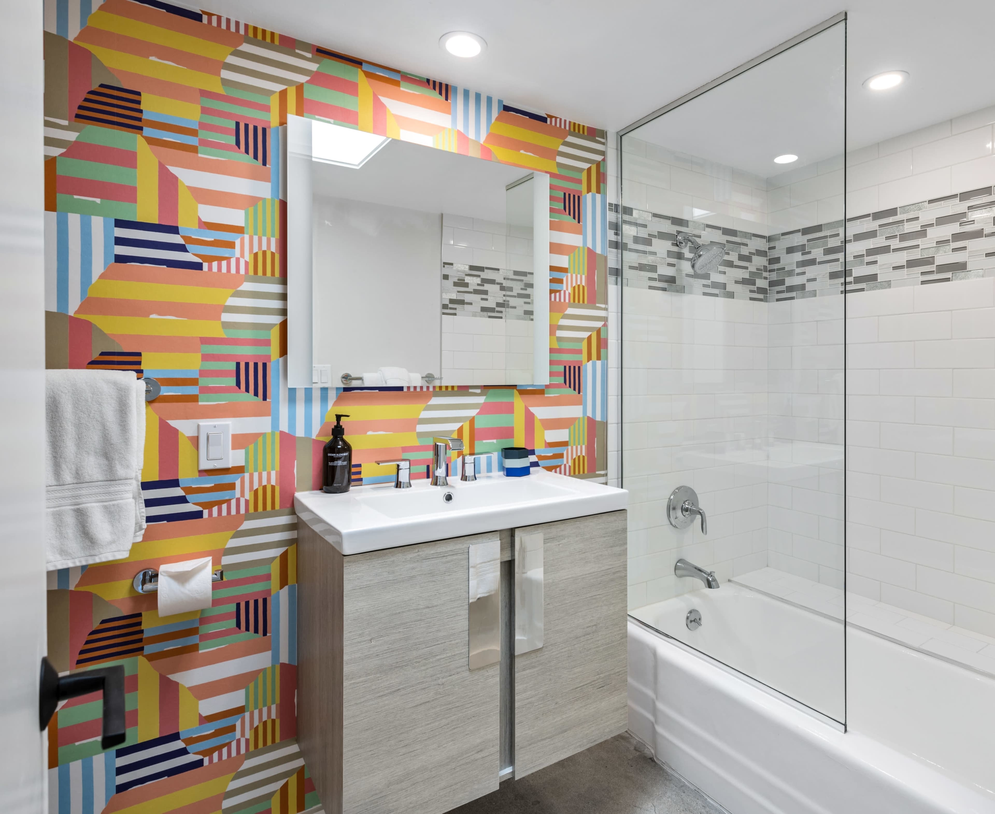 A brightly decorated bathroom features a patterned wallpaper, a modern sink with a mirror above it, and a glass shower enclosure beside a bathtub.