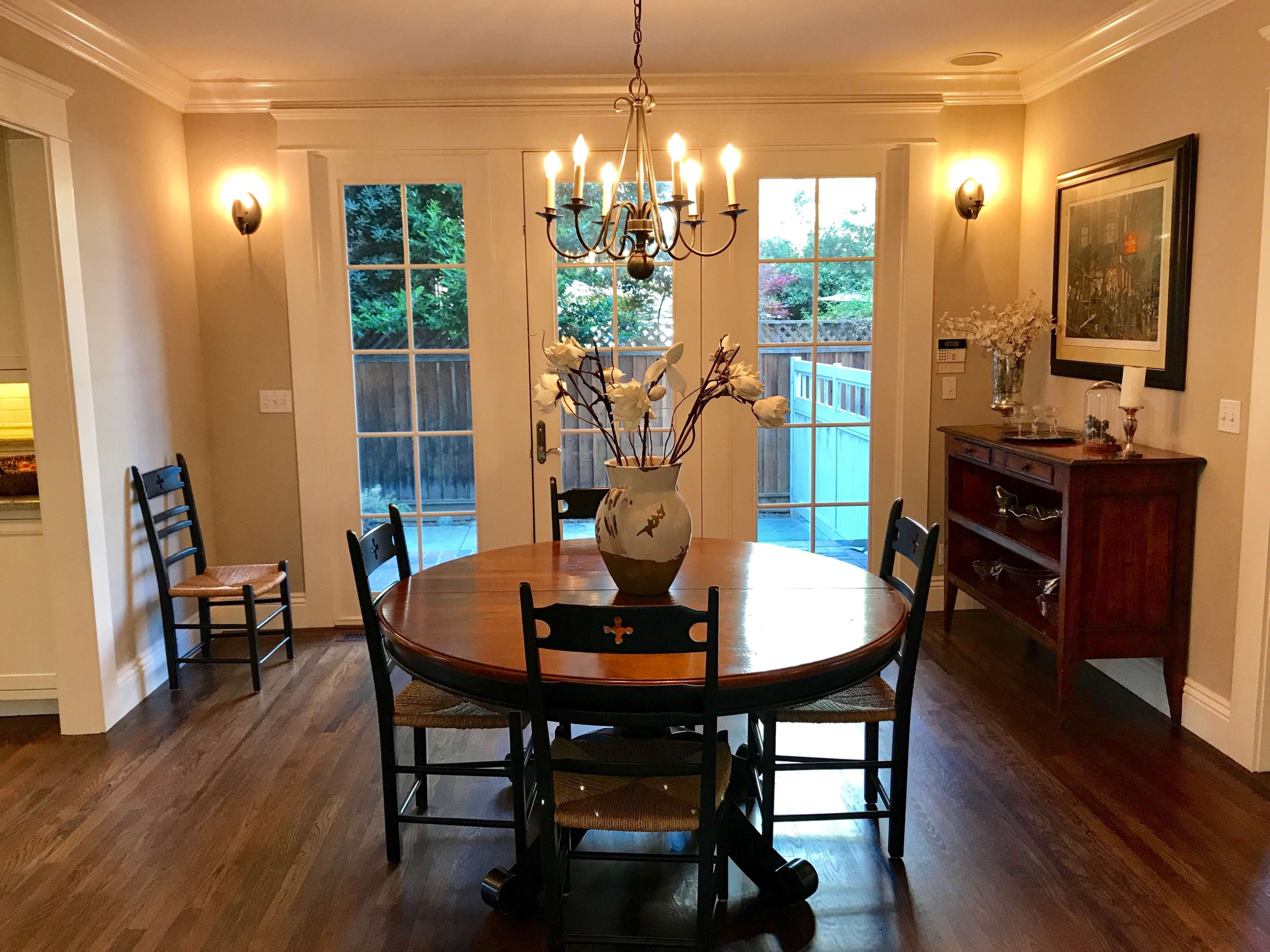 The image shows a dining area with a round wooden table surrounded by chairs, a vase of flowers in the center, and a chandelier overhead, facing a set of glass doors.
