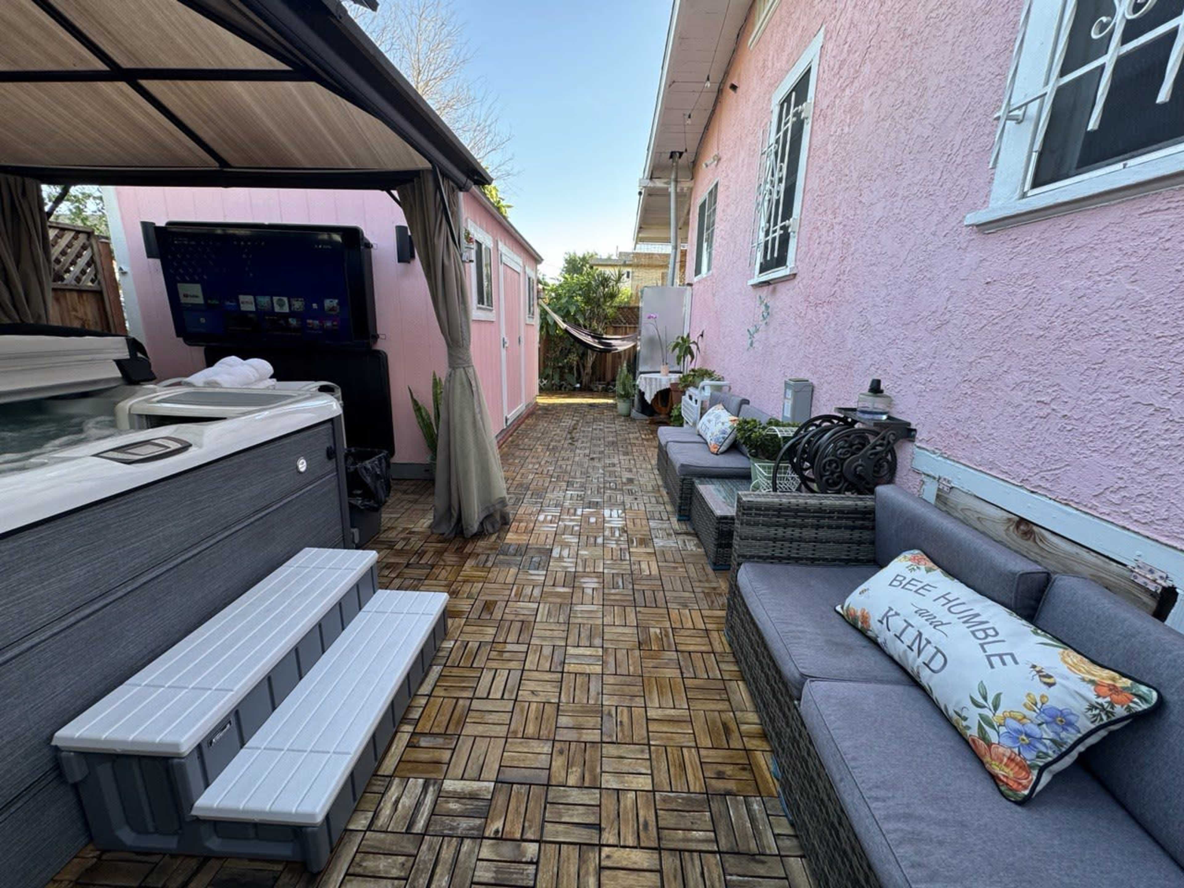 A backyard area with a hot tub, seating arrangements, and a walkway, bordered by a pink wall and greenery.