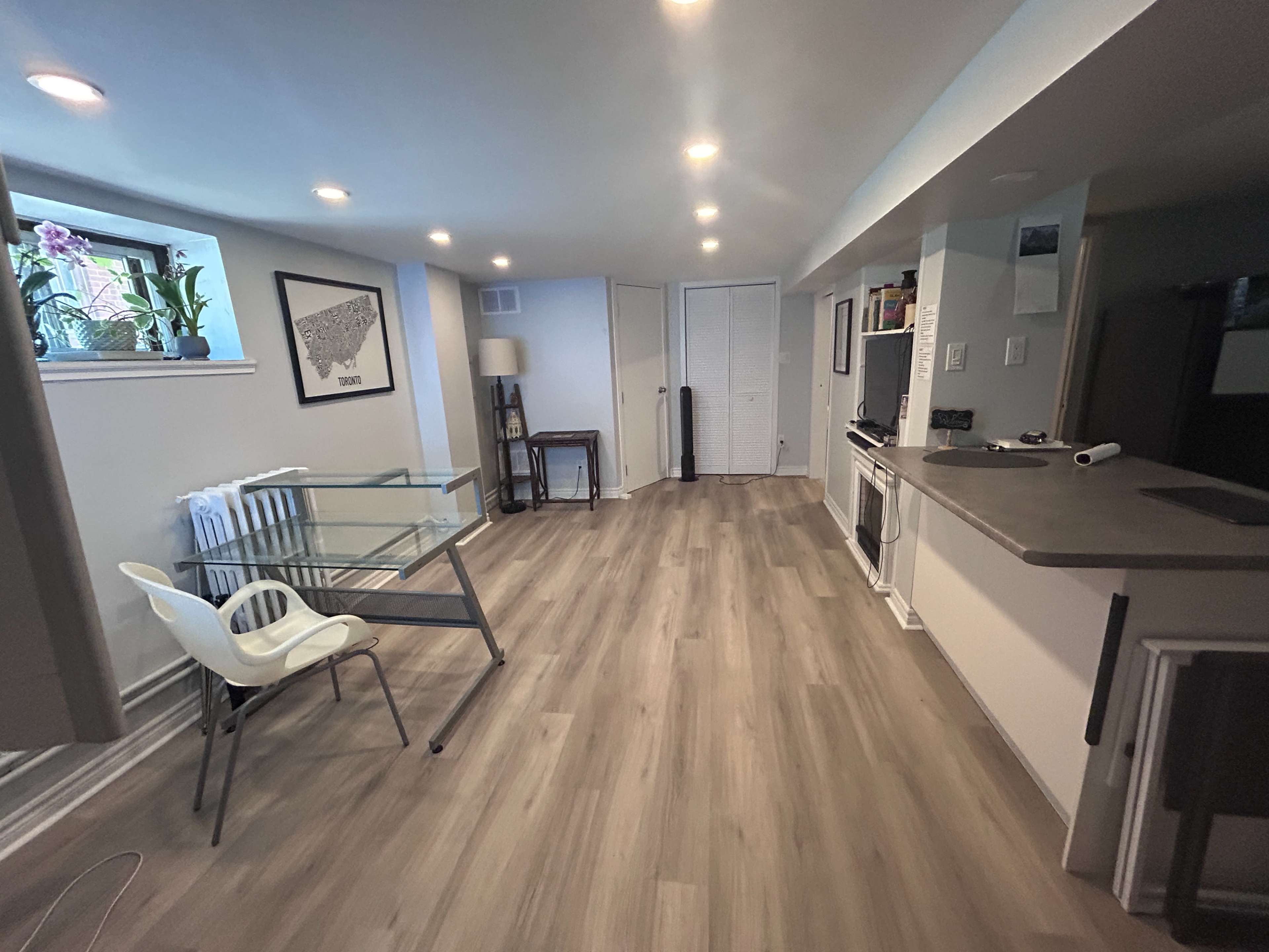 The image shows a spacious, well-lit room with hardwood flooring, featuring a glass table, a chair, a lamp, and a kitchenette area.