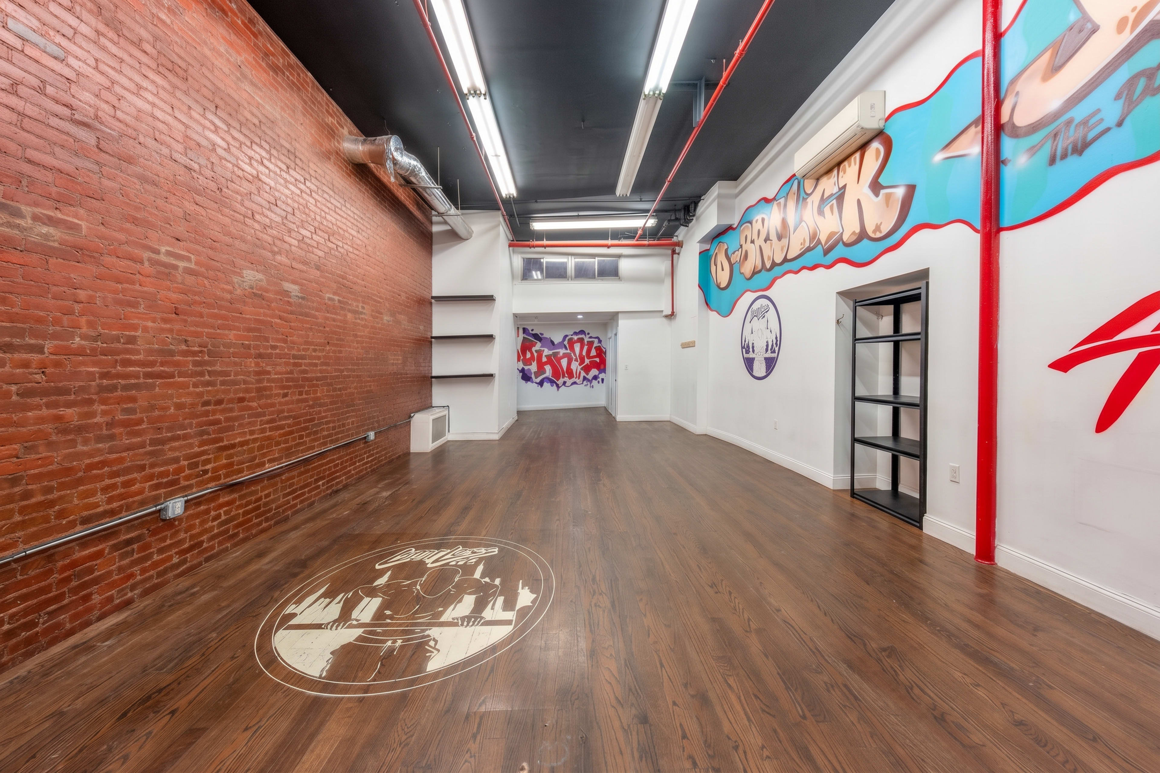 The image shows a spacious room with exposed brick walls, hardwood flooring, and colorful graffiti-style murals.
