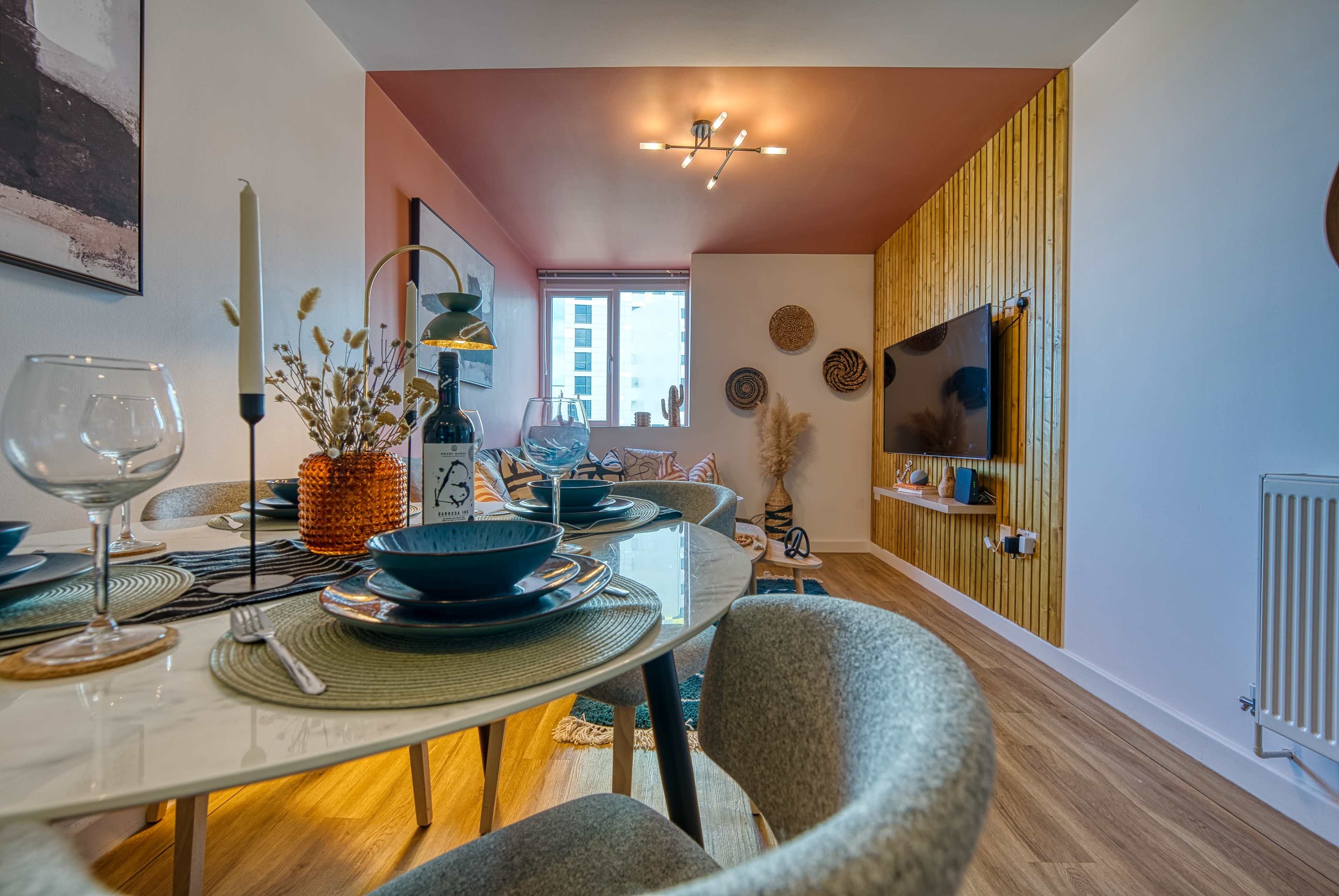 A modern dining area with a round table set for a meal, leading into a cozy living space featuring a television and decorative wall elements.