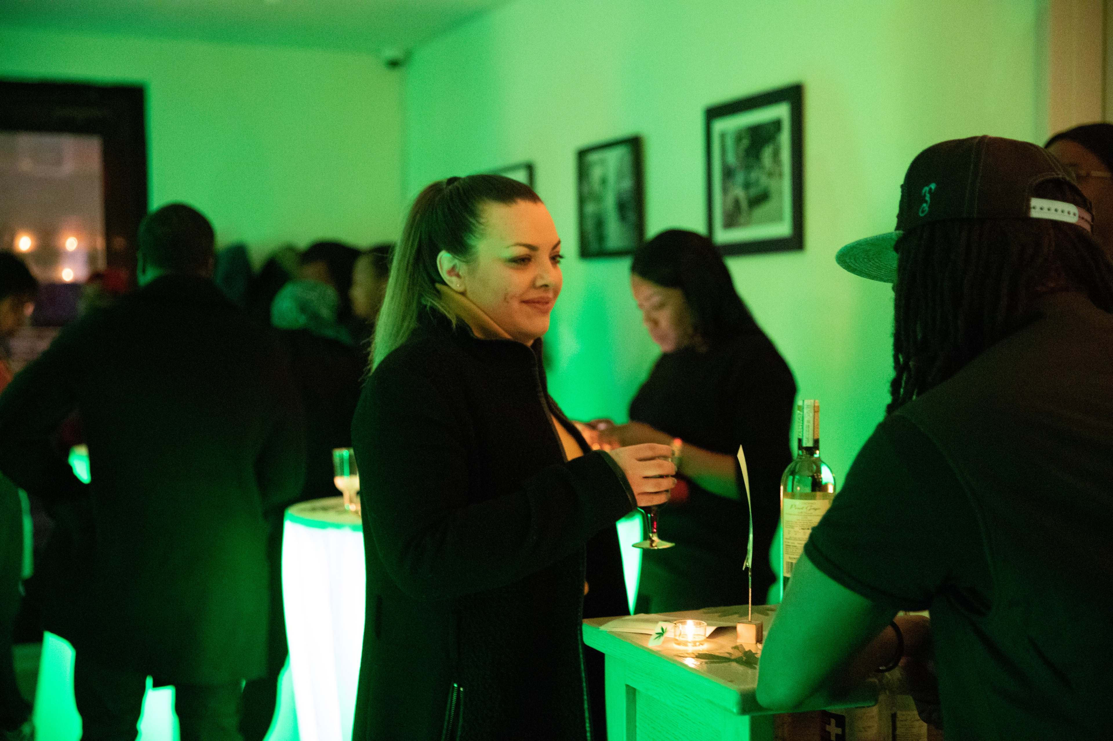 A group of people socialize in a dimly lit bar with green lighting while holding drinks.