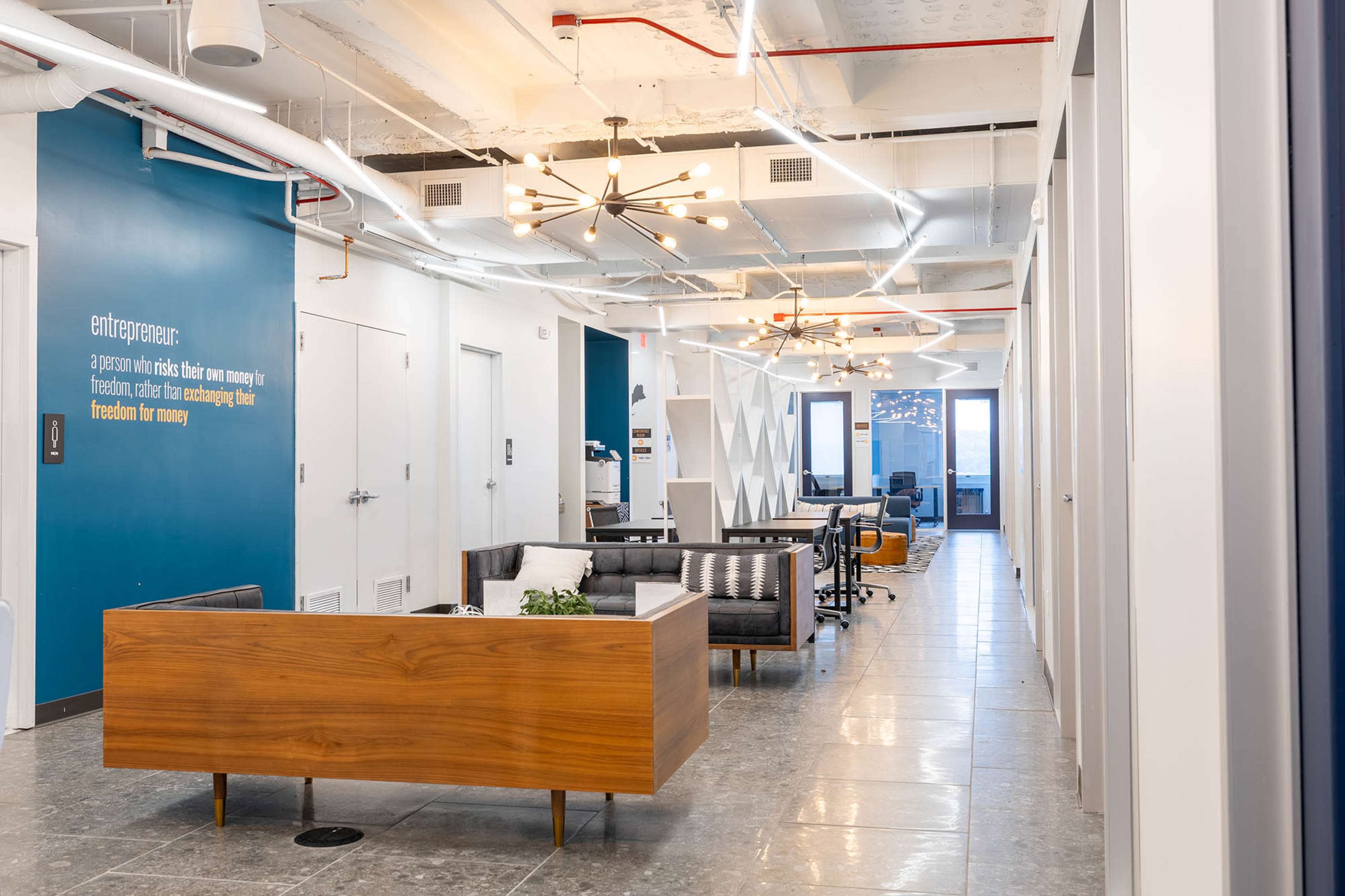 The image shows a modern office interior featuring a reception area with a wooden desk, a seating arrangement, and workspaces arranged along the hallway.