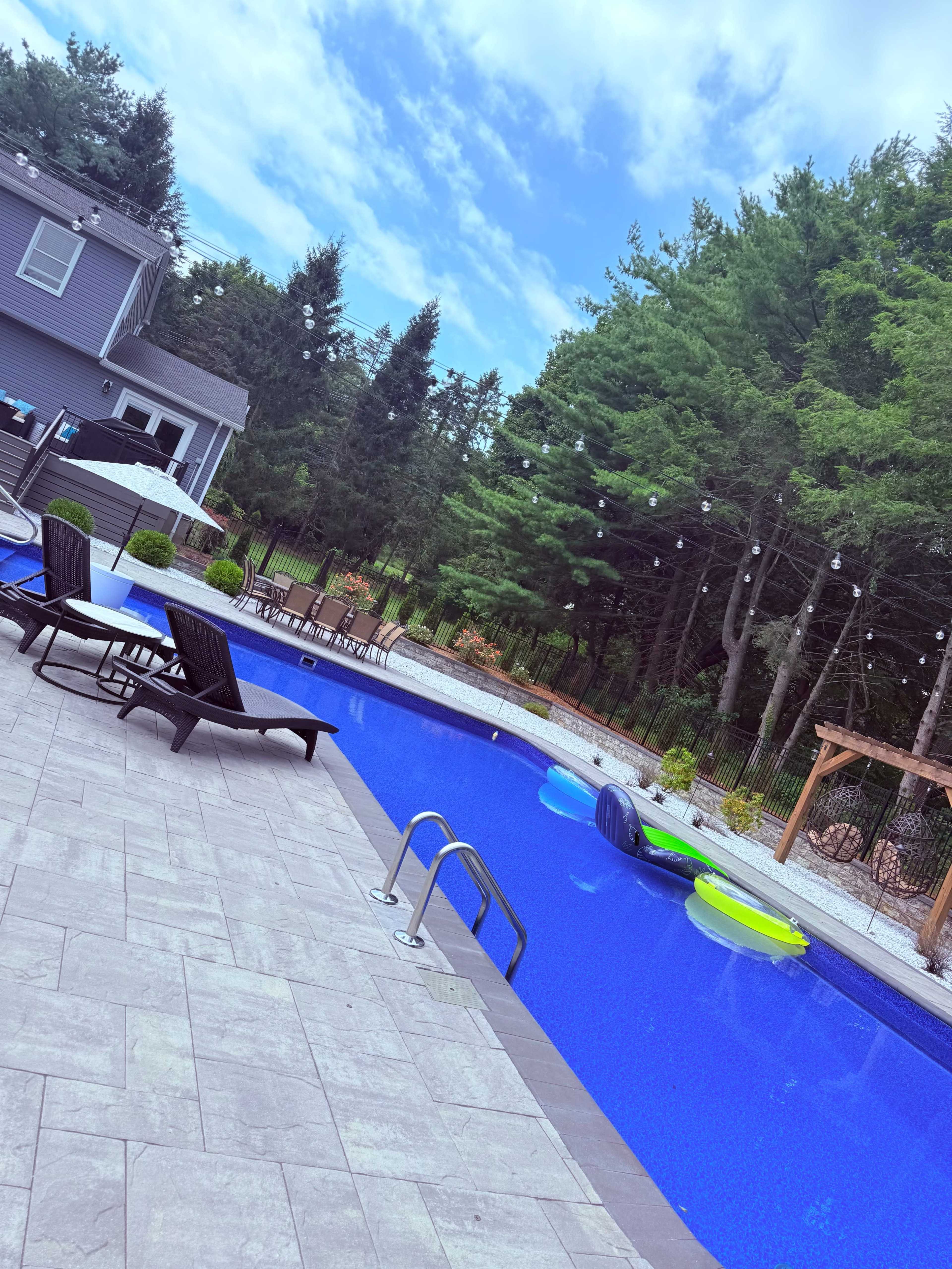 The image shows a rectangular swimming pool surrounded by lounge chairs and colorful inflatable floats, with a backdrop of trees and a house.