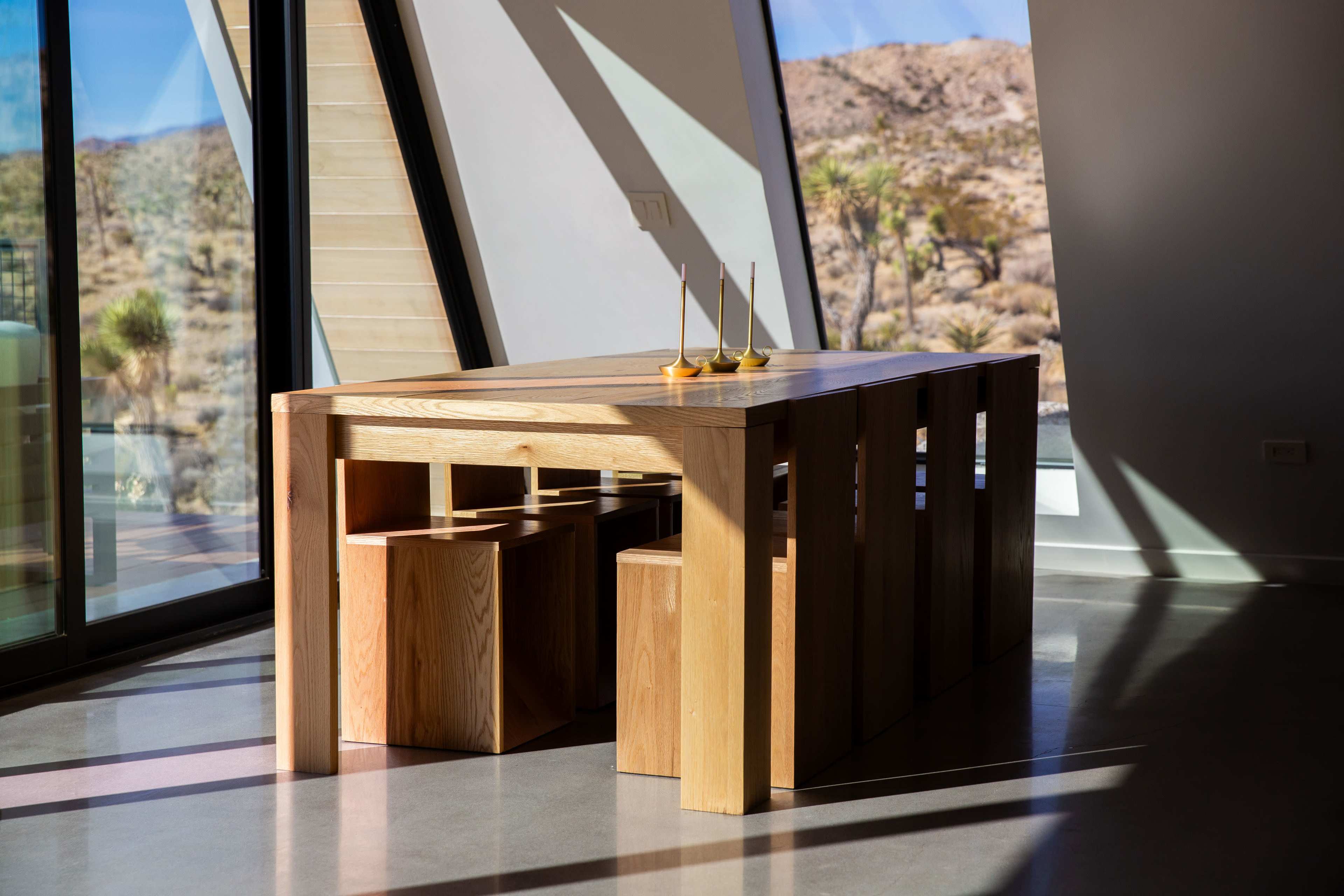 A wooden dining table with block stools is positioned near large windows that provide a view of a desert landscape.