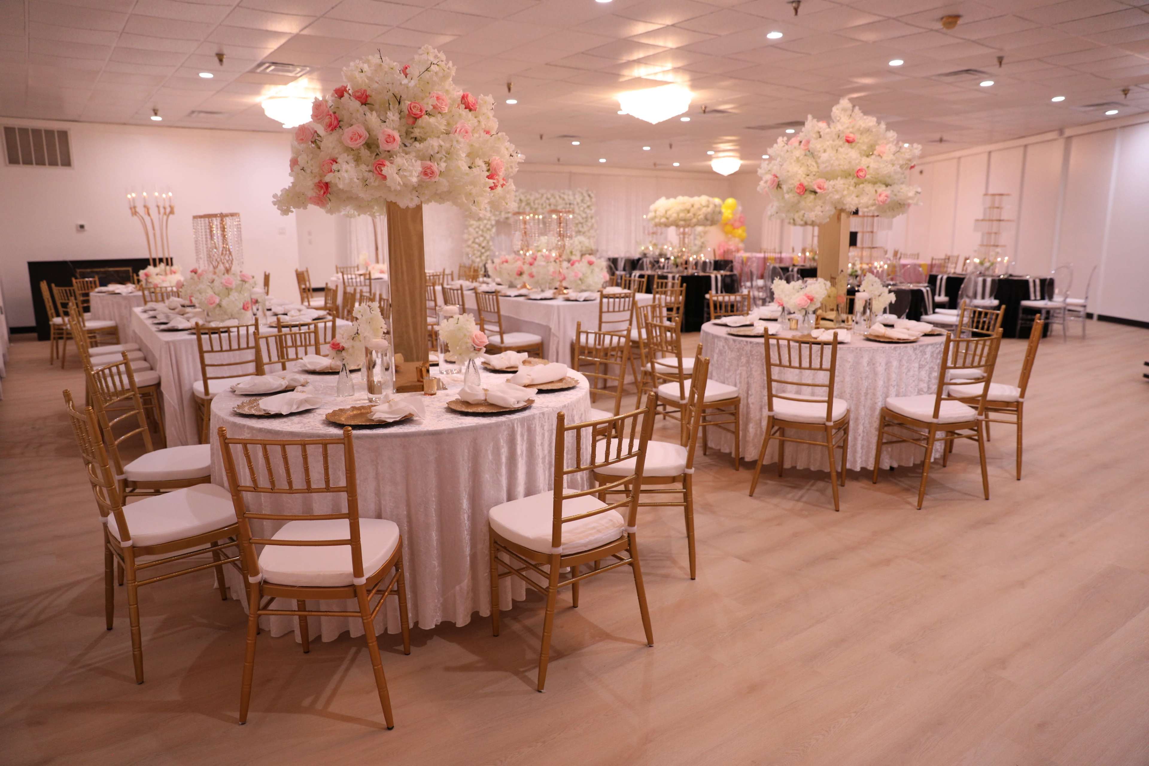 A spacious banquet hall features elegantly set round tables adorned with white tablecloths, gold chargers, and large floral centerpieces.