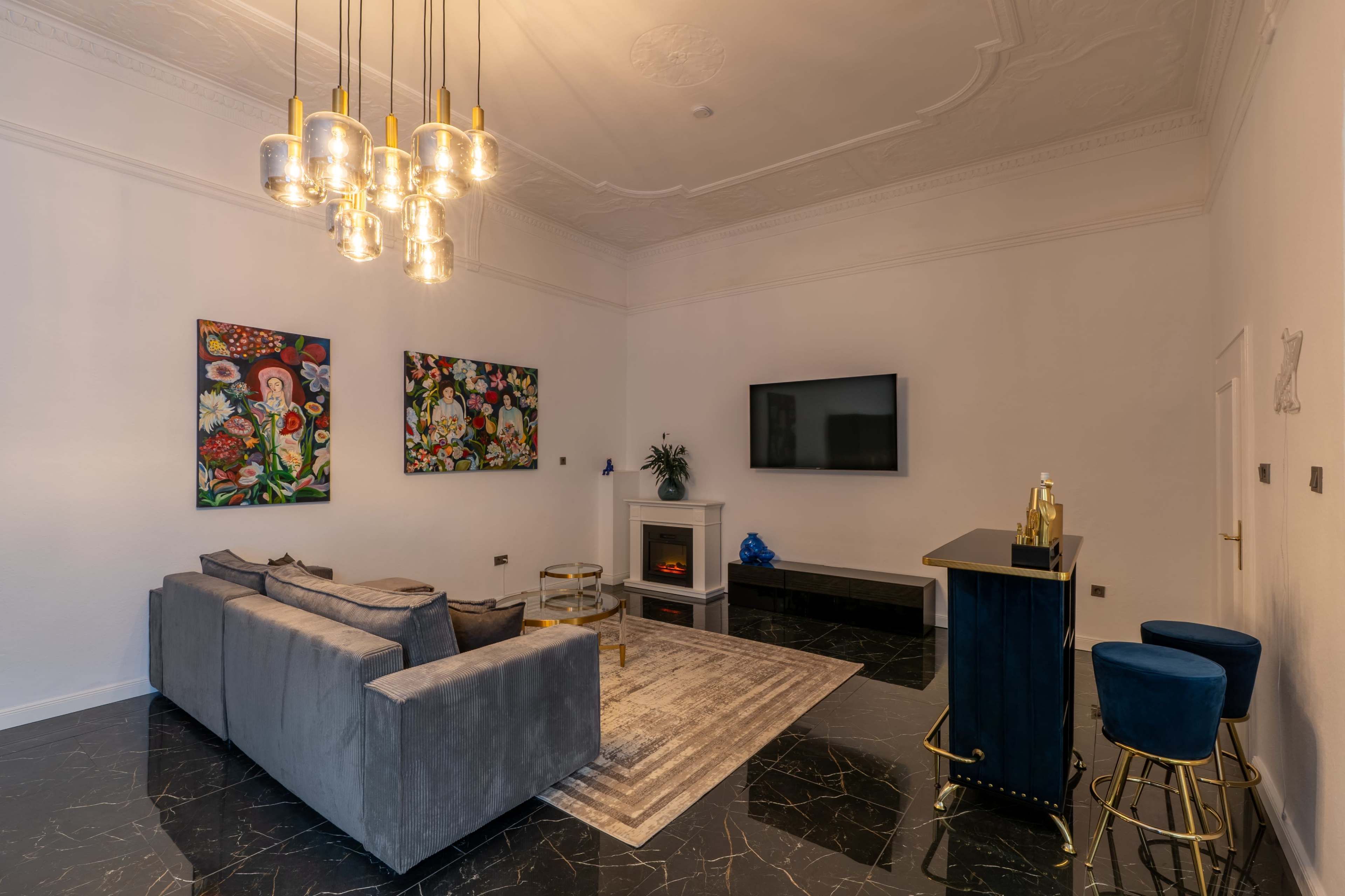 The scene features a modern living room with a gray sofa, colorful flower paintings on the walls, a sleek bar counter, and a television mounted above a fireplace.