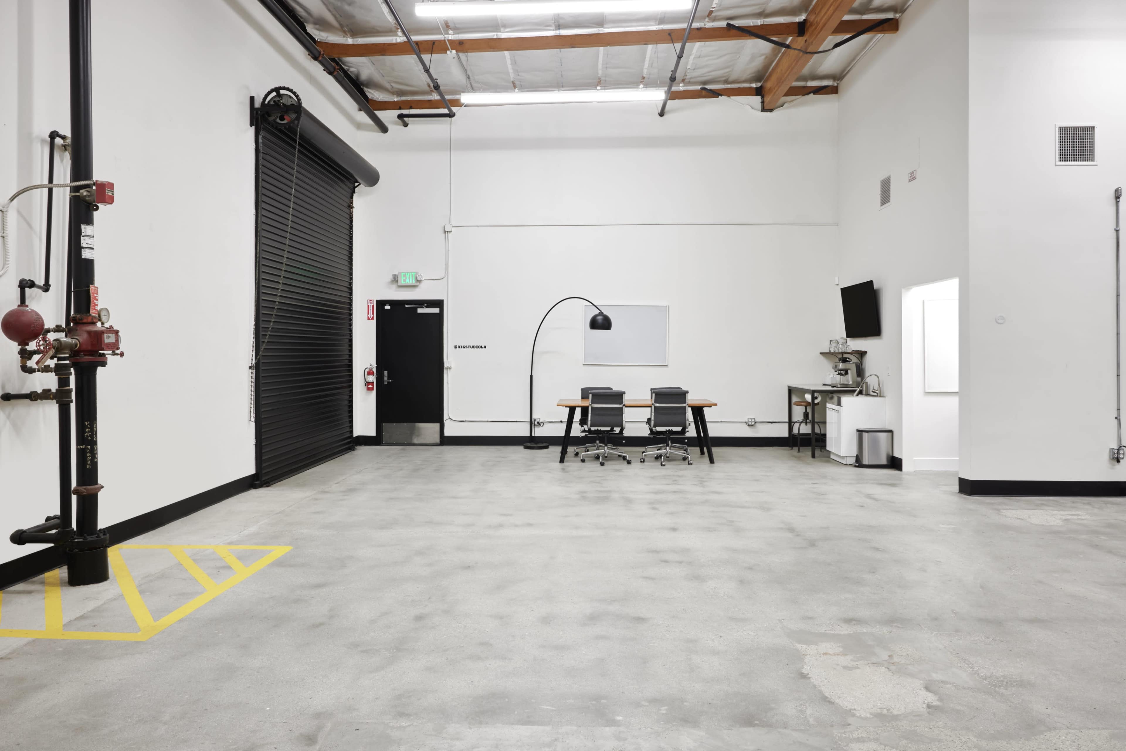 The image depicts an empty industrial space featuring a conference table with chairs, a wall-mounted monitor, and a large black roll-up door.