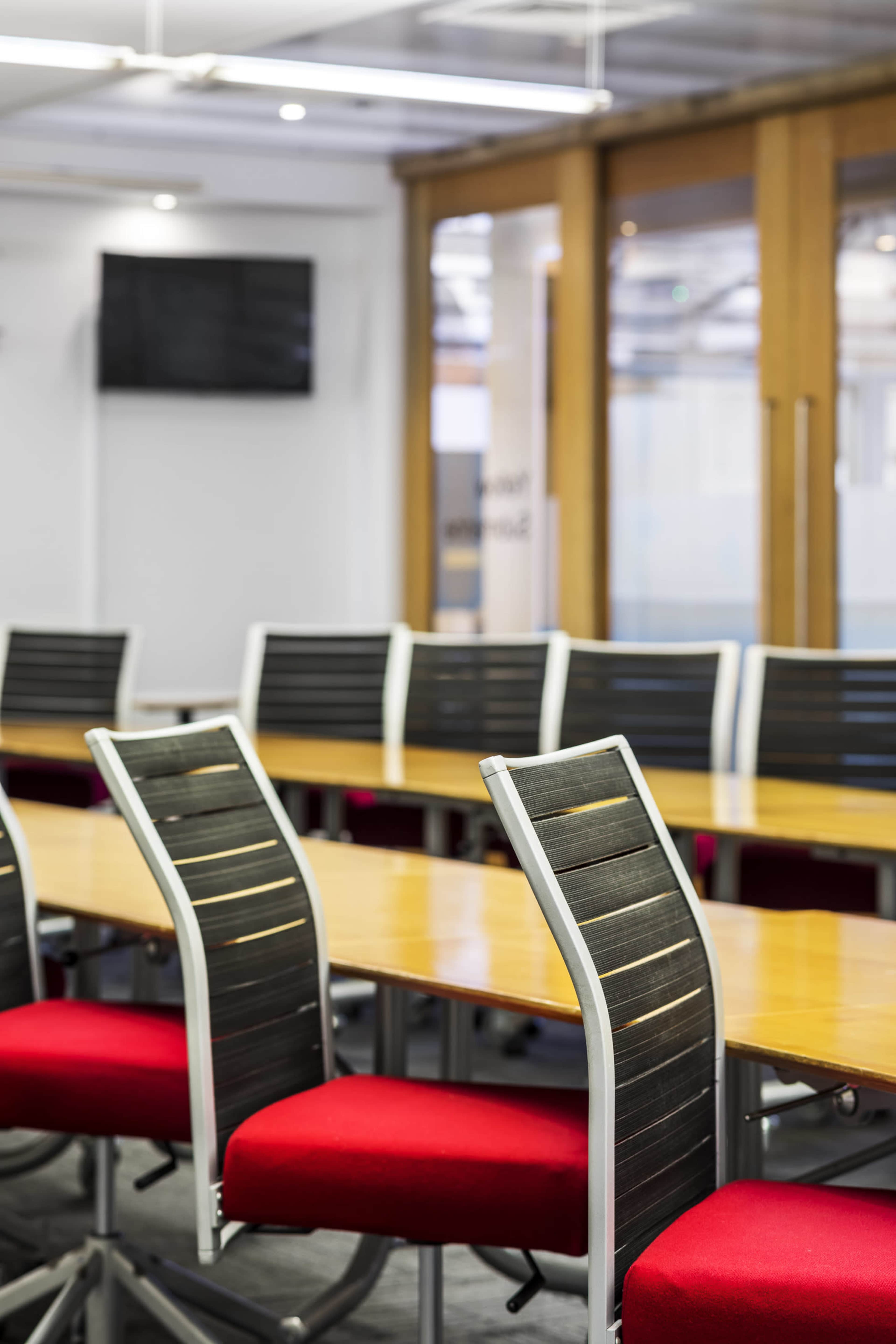 A conference room features several long tables with red upholstered chairs and a television mounted on the wall.