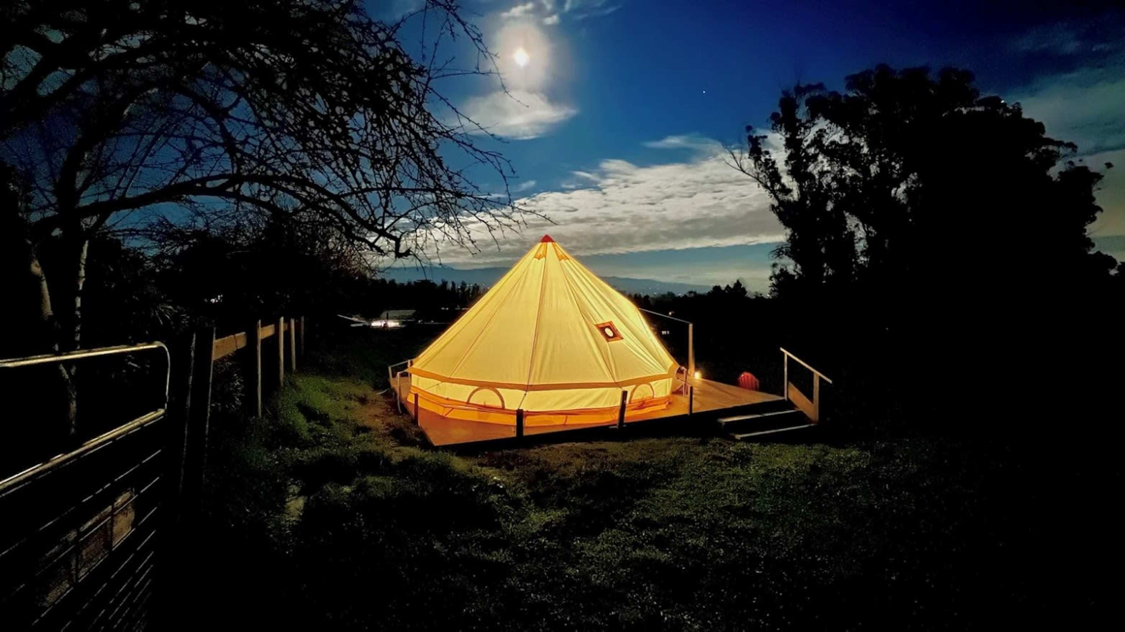 A illuminated tent stands on a wooden platform under a moonlit sky, surrounded by trees and clouds.
