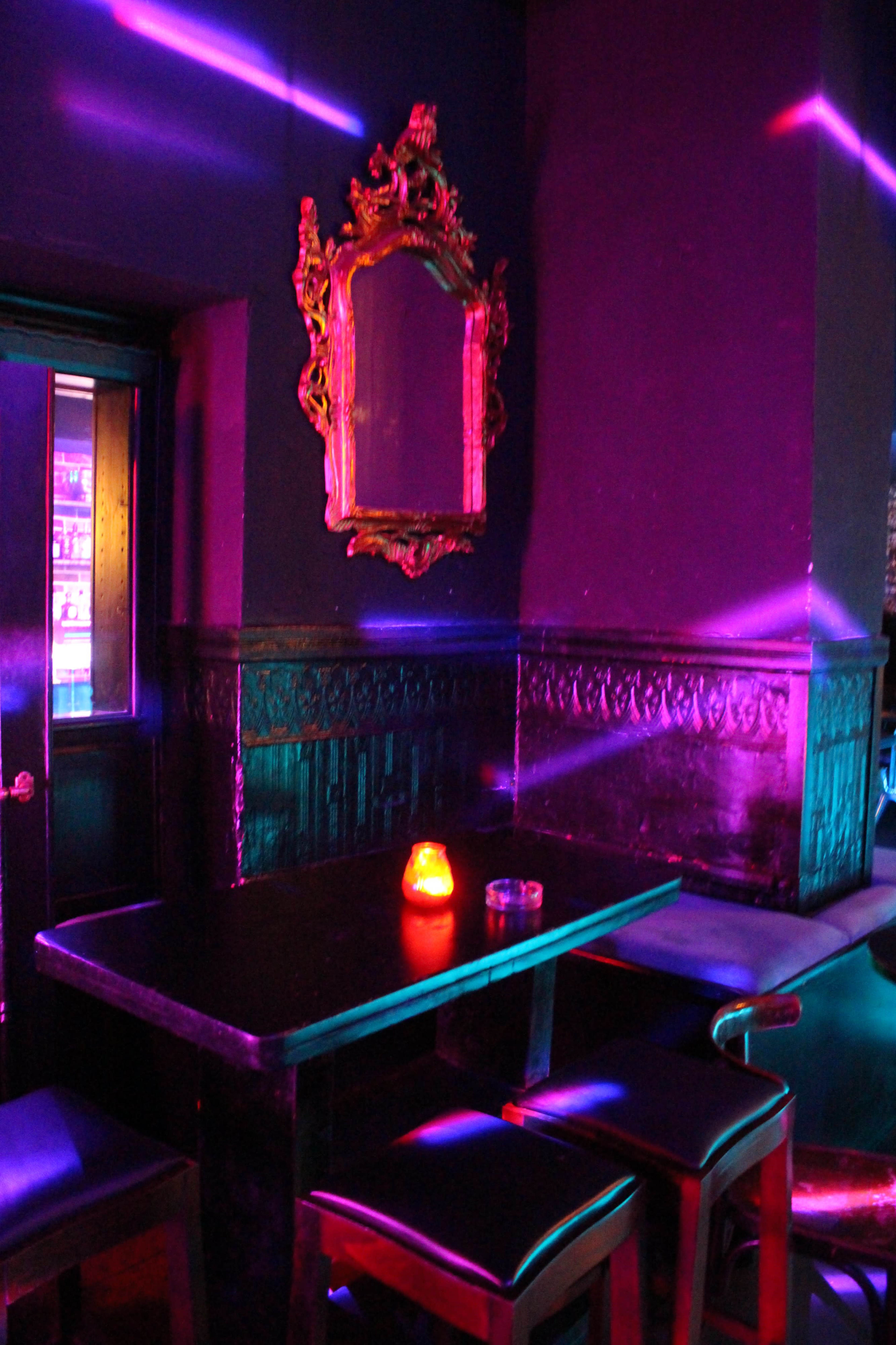 The image shows a dimly lit bar with a dark wooden table and chairs, a glowing candle on the table, and a wall-mounted mirror reflecting colored lights.