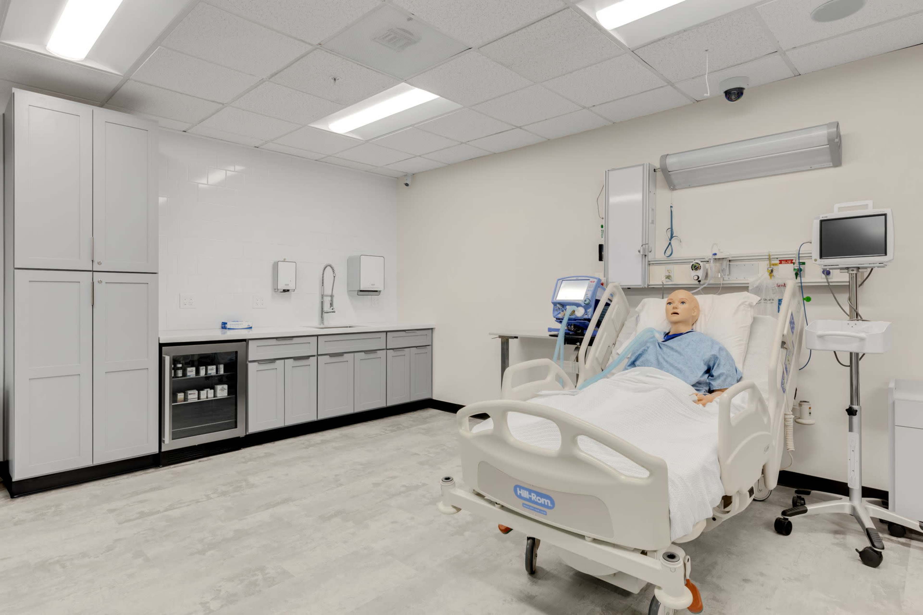 A hospital room equipped with medical equipment, a mannequin on a hospital bed, and storage cabinets along the wall.