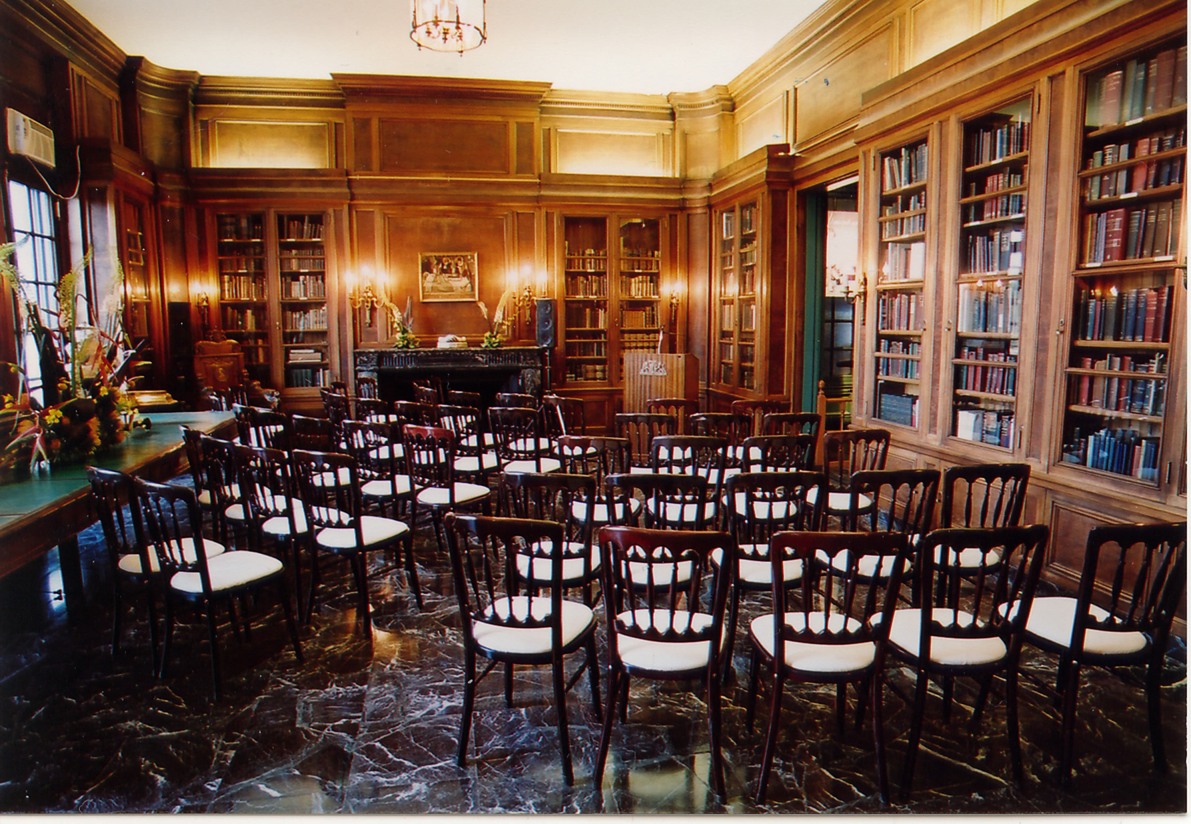 The image shows a library room with wooden paneling, a piano, and rows of wooden chairs arranged for an event.