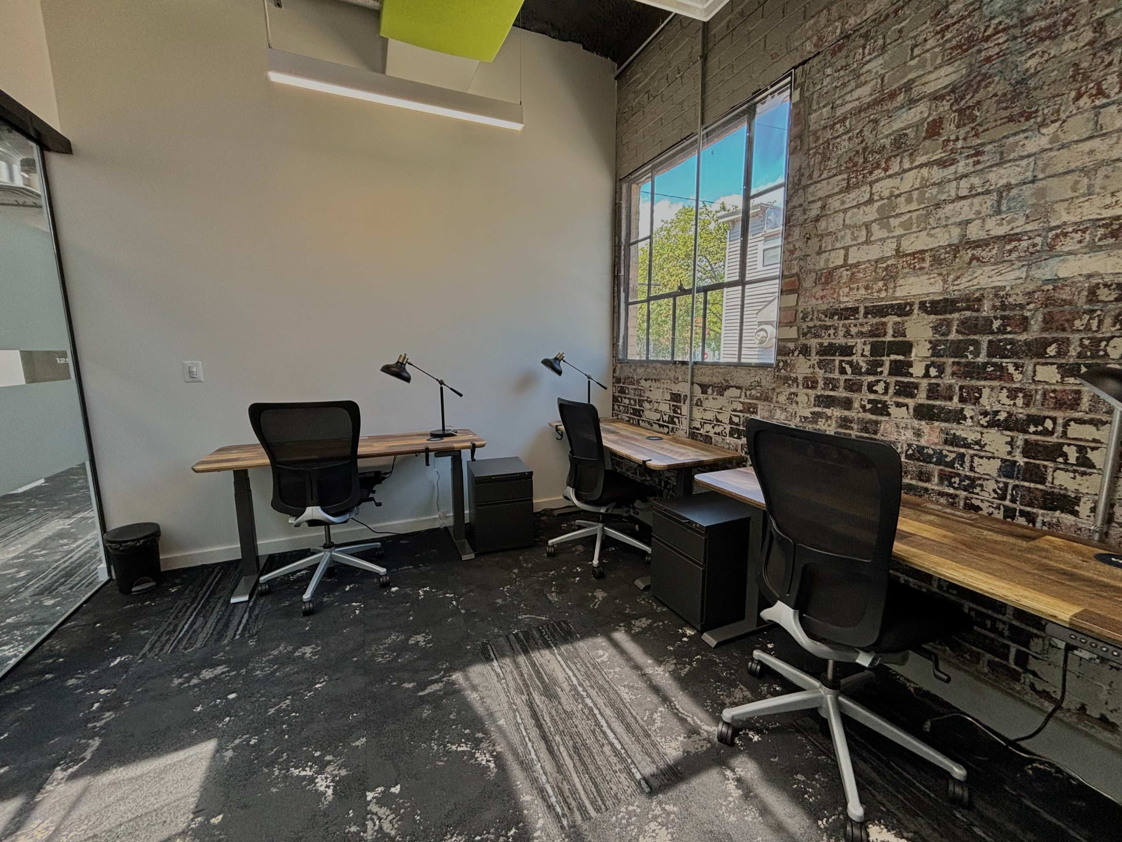A small office space with two wooden desks, black office chairs, and large windows allowing natural light to illuminate the brick walls.
