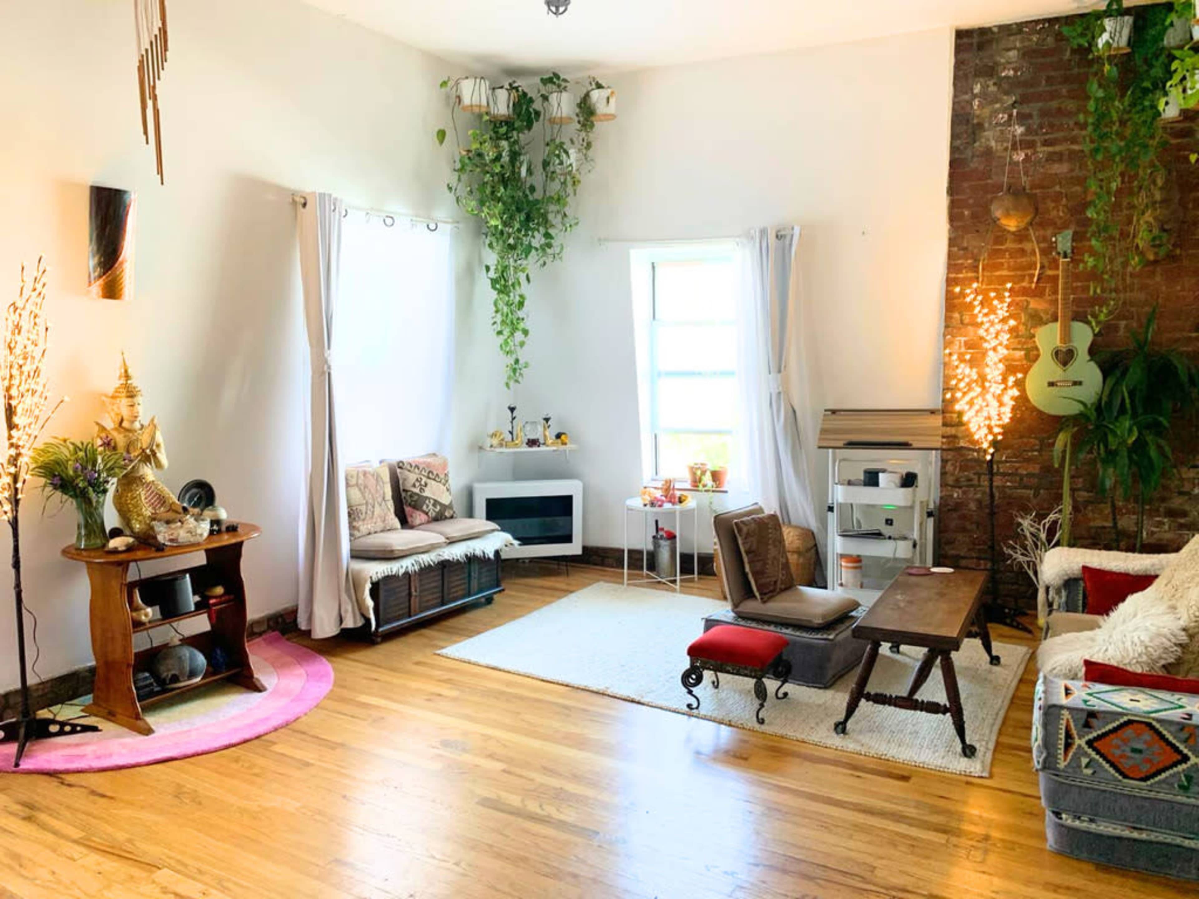 The room features hardwood floors, a mix of seating options, plants hanging from the ceiling, and a brick wall with decorative elements.
