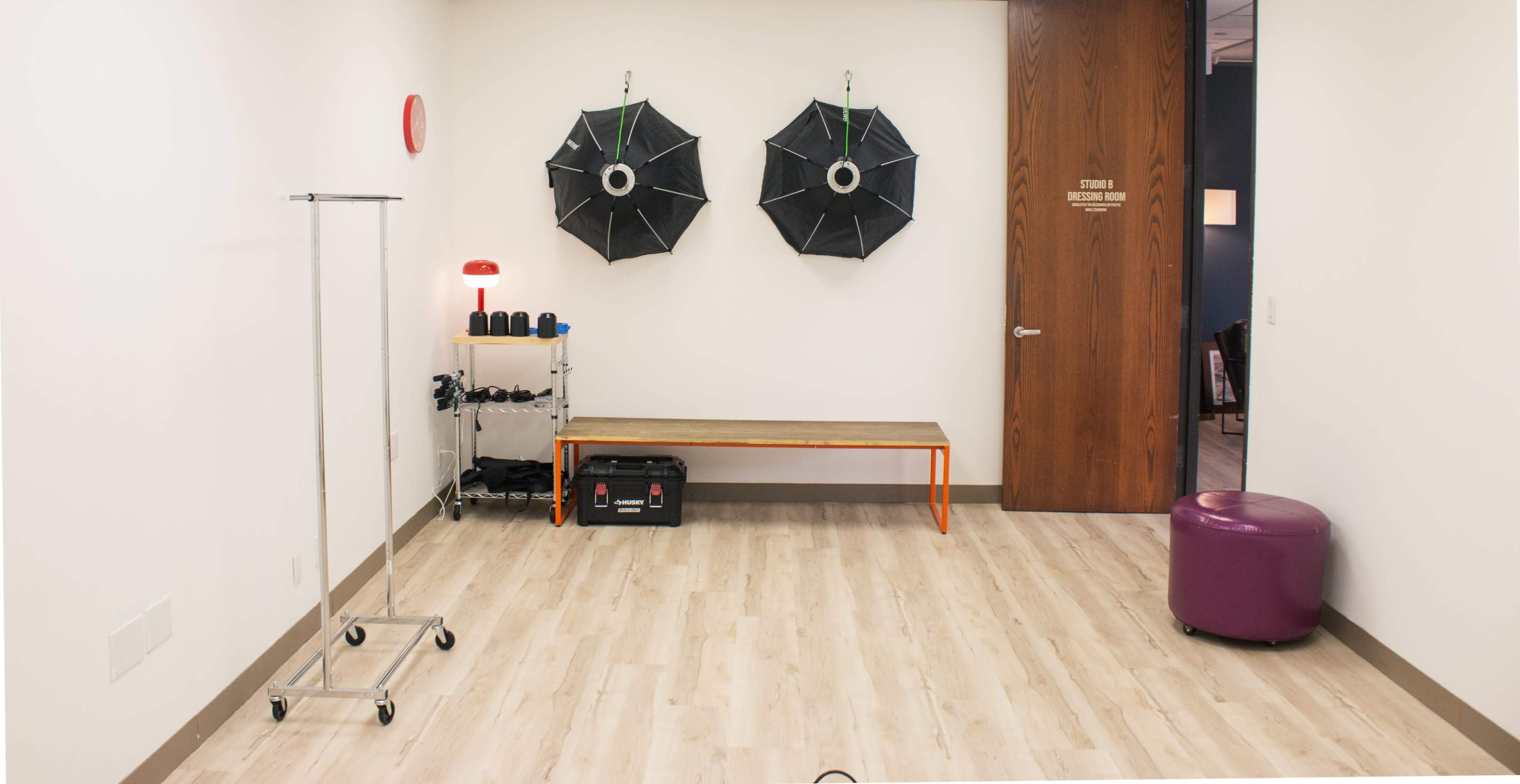 The image shows a sparsely furnished room featuring two black umbrellas mounted on the wall, a wooden bench, a rolling cart with weights, and a circular purple stool.