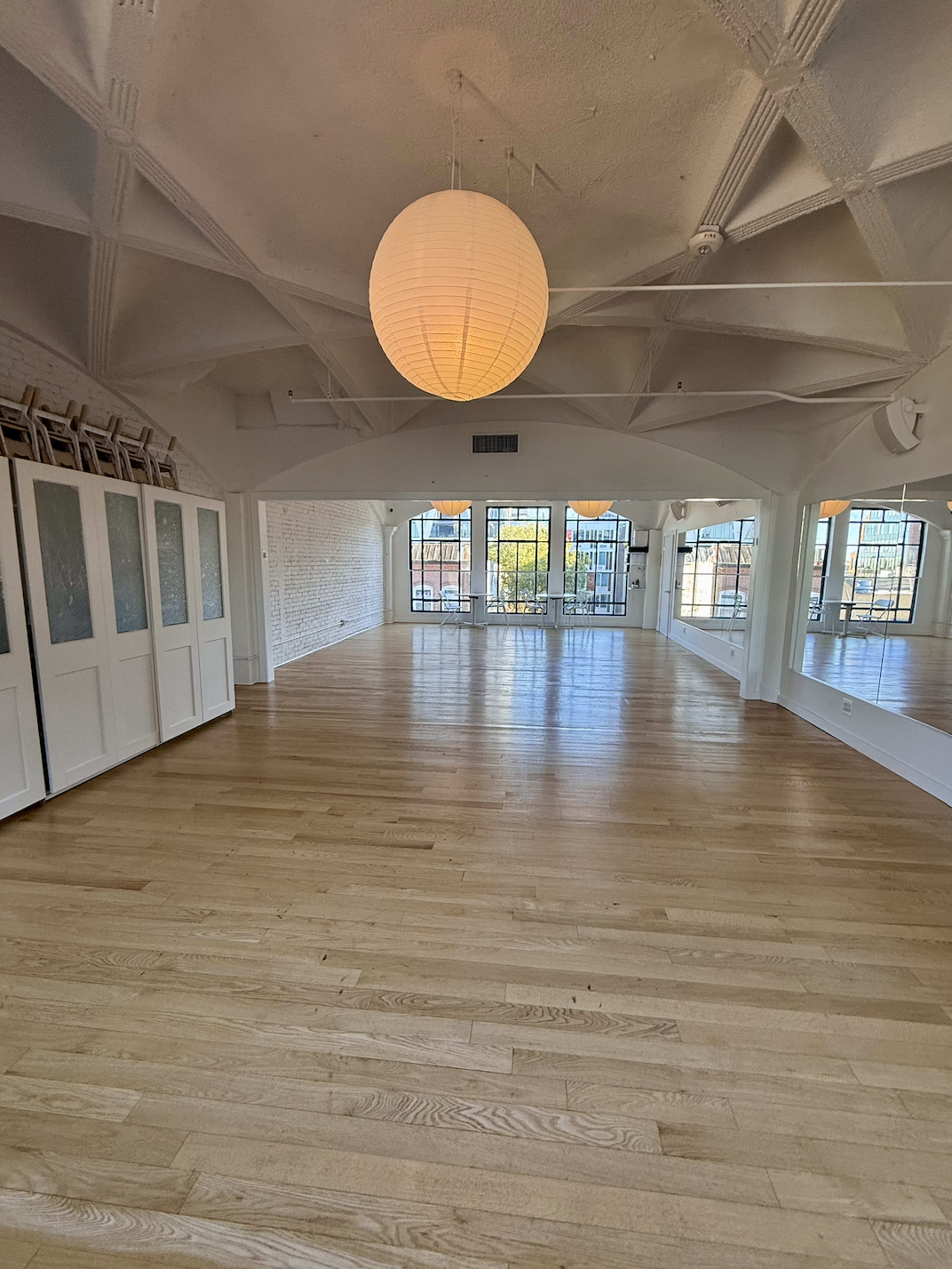 The image shows a spacious, well-lit room with wooden floors, large windows, and circular pendant lights hanging from a high ceiling.