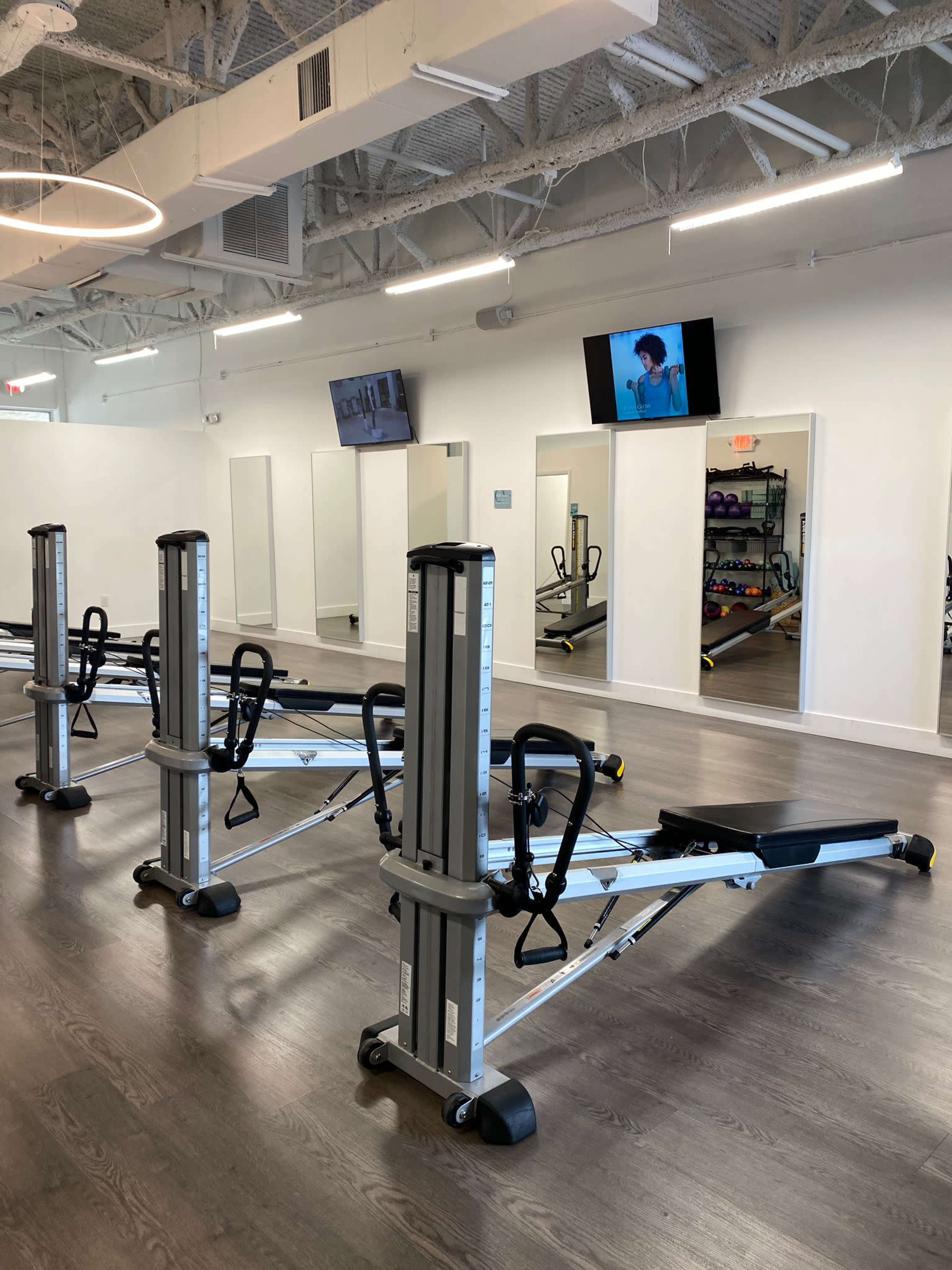 The image shows a modern fitness studio with several rowing machines aligned in a well-lit space featuring mirrors and wall-mounted screens.
