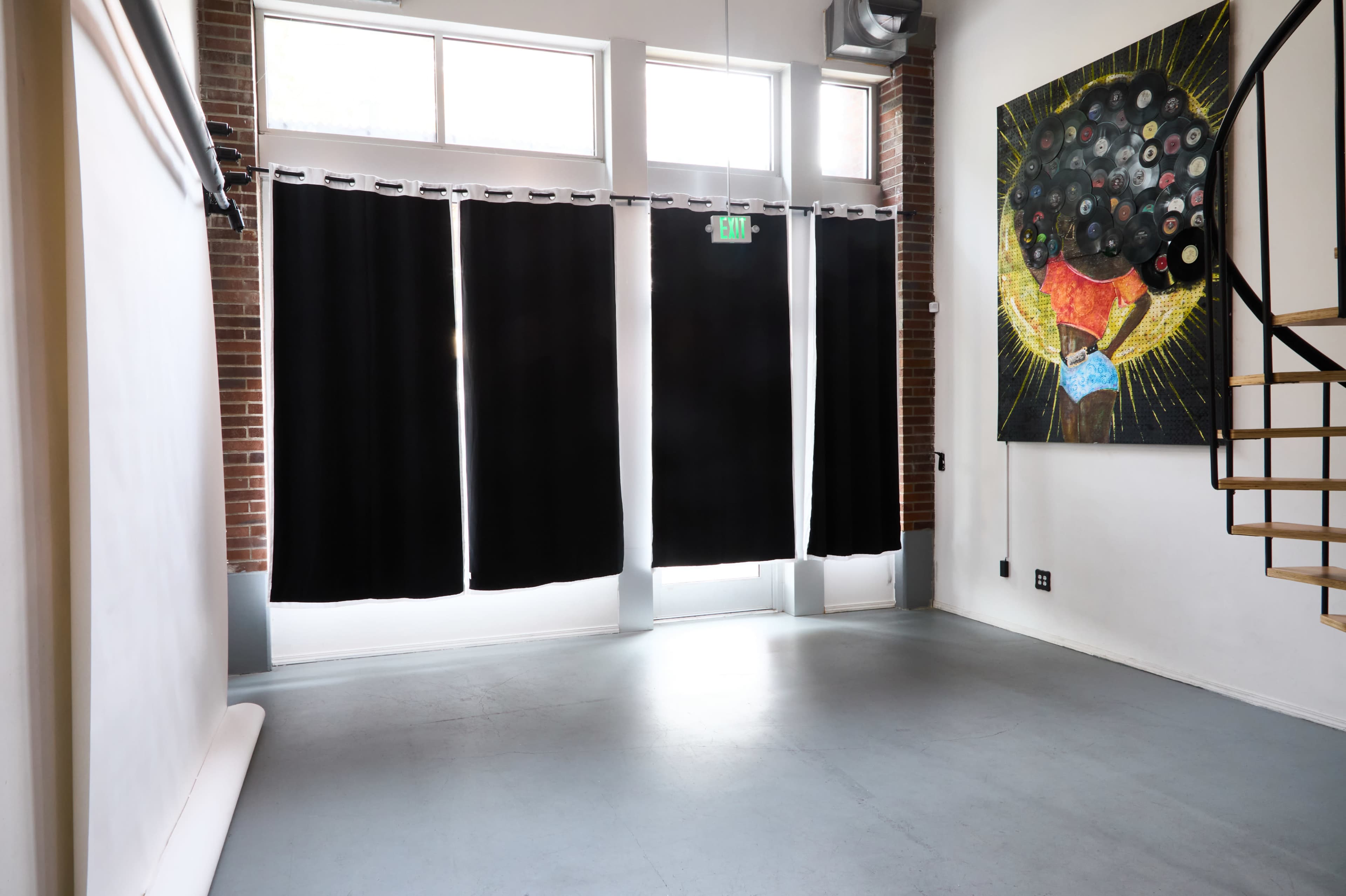 The image shows a spacious interior room with black curtains covering large windows and a colorful artwork featuring a figure surrounded by records on one wall.