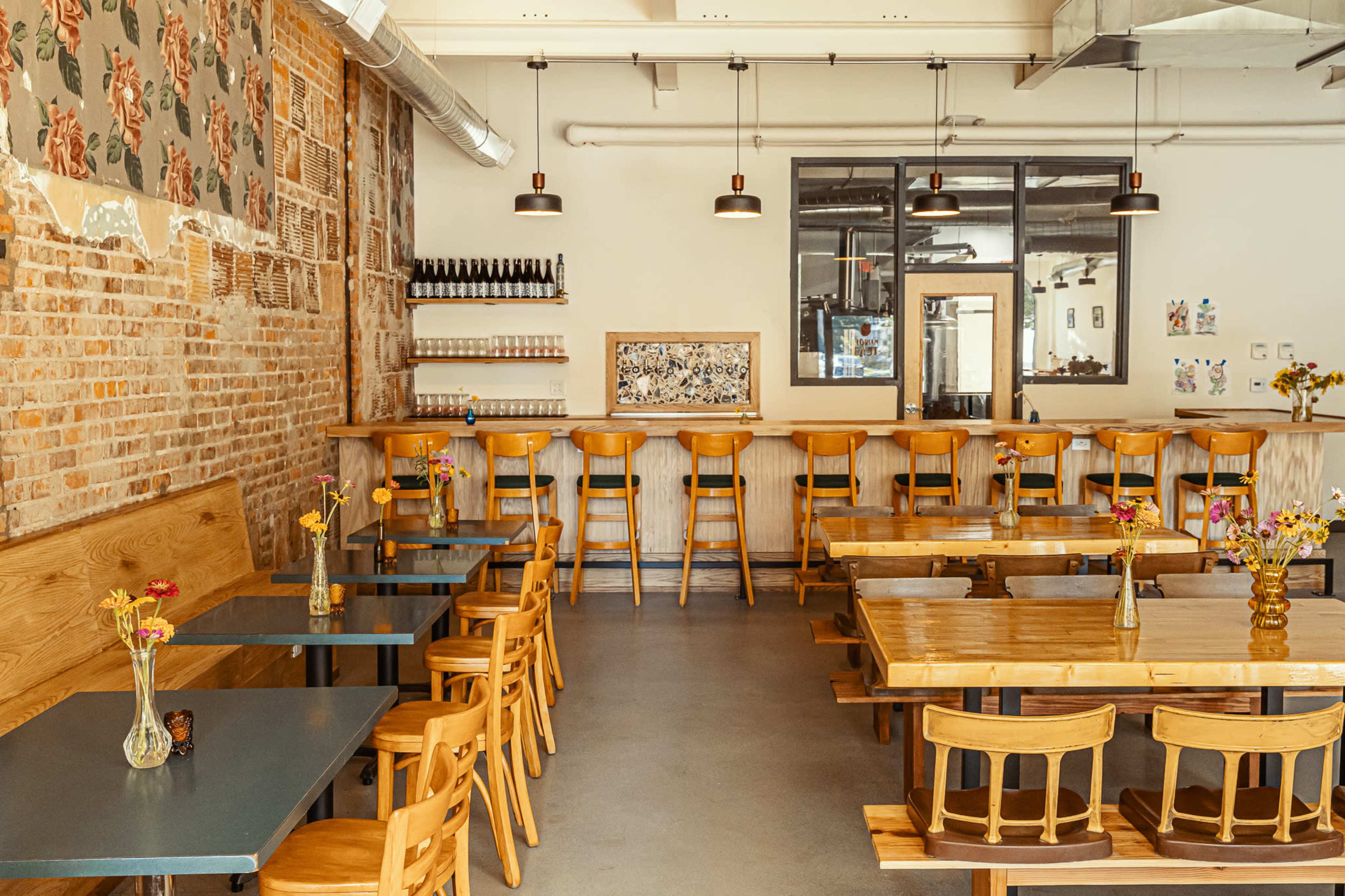 The image shows a casual restaurant interior with wooden tables, chairs, and a bar area lined with high stools, featuring decorative flowers on the tables and a brick wall backdrop.