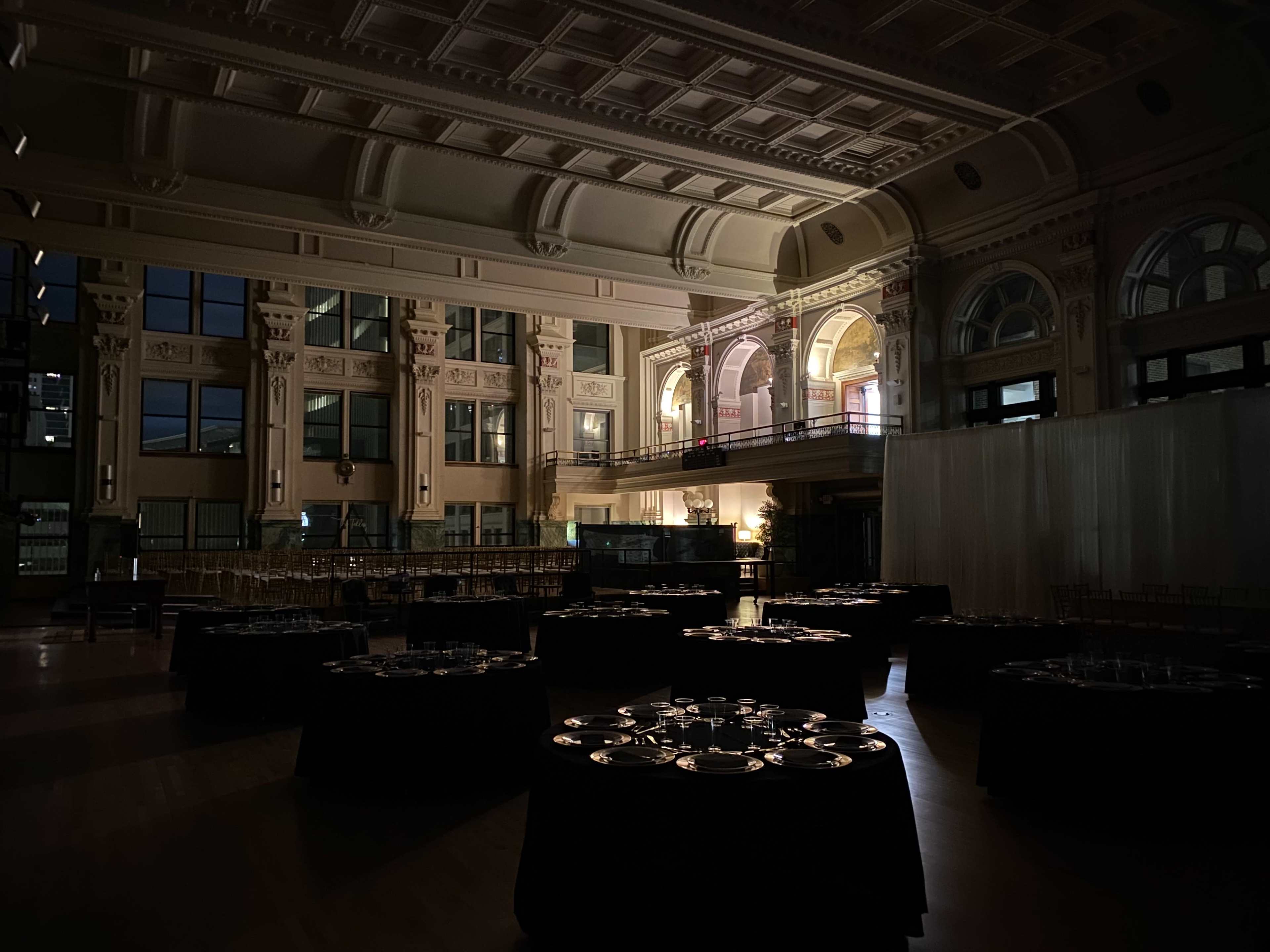 A dimly lit event space with round tables set for a formal dinner, featuring high ceilings and architectural details.