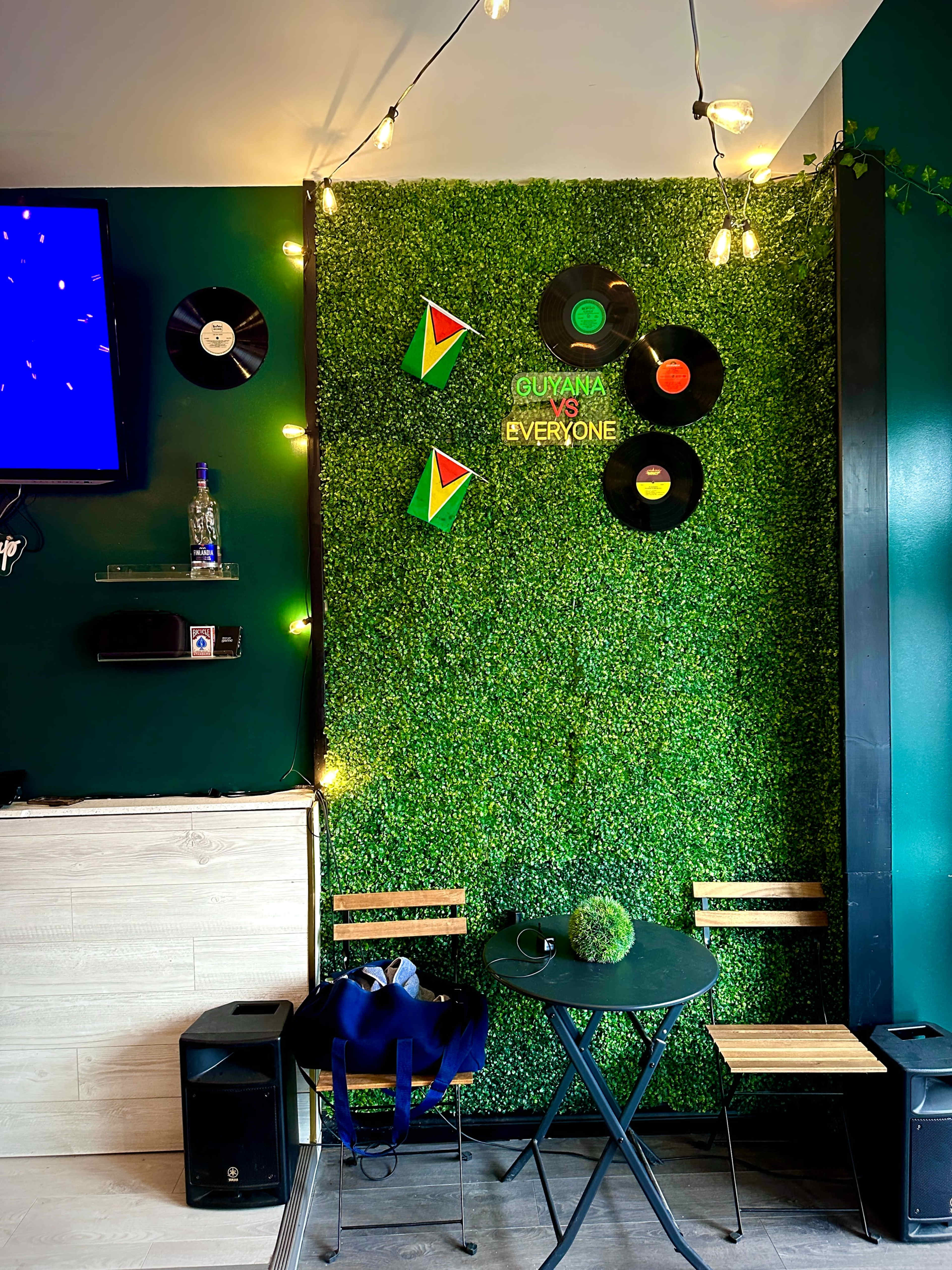 The image shows a small café corner with a green wall adorned with vinyl records and geometric decorations, alongside a round table and two wooden chairs.