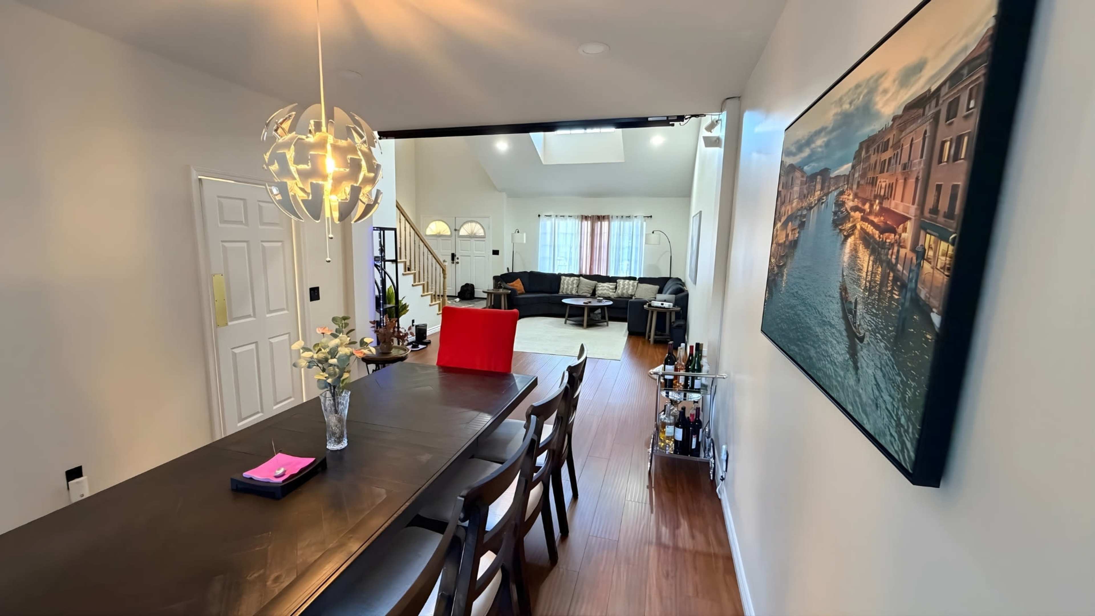 A dining area with a dark table and chairs leads to a living room featuring a large window and a view of a picturesque landscape.