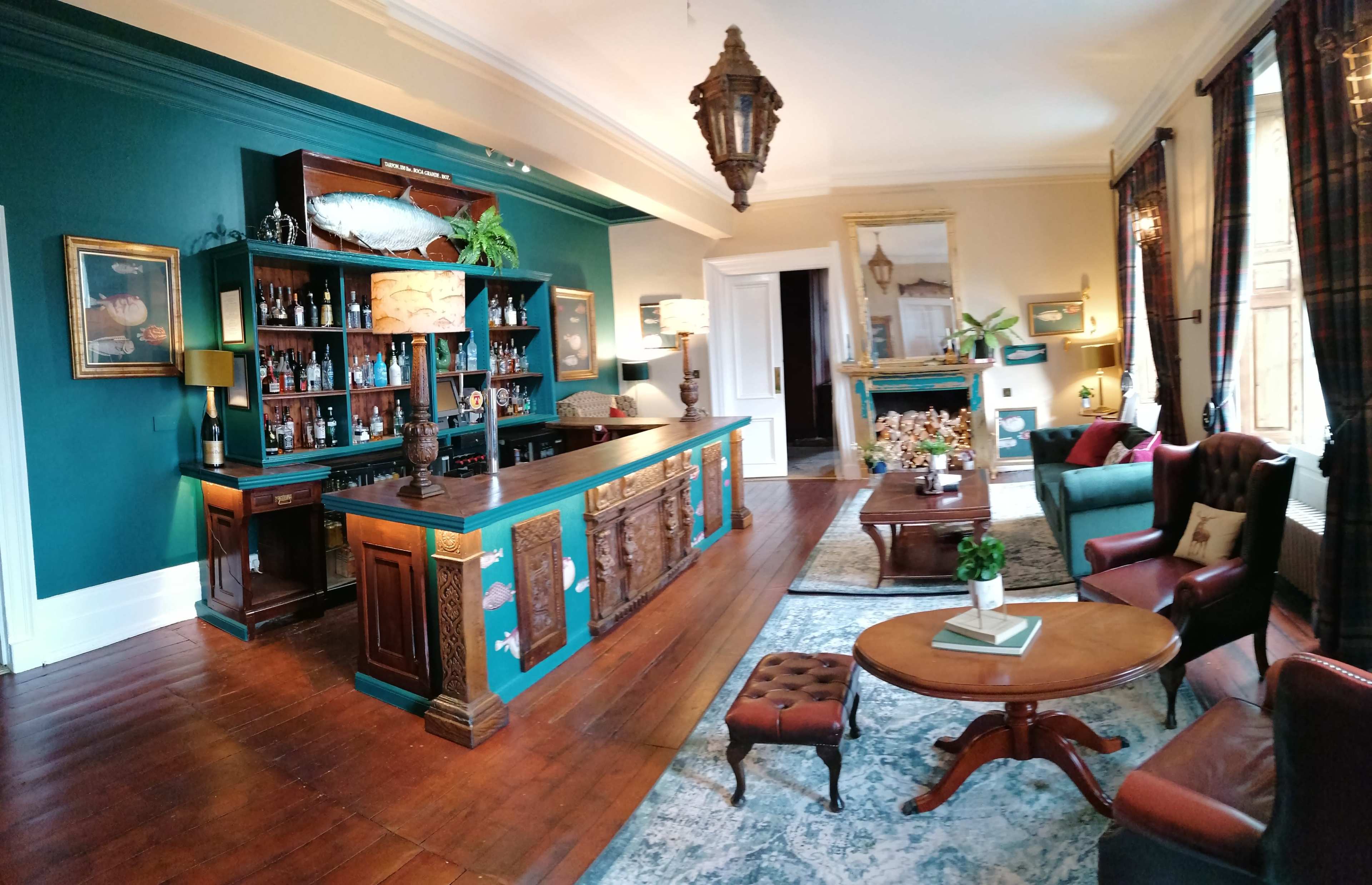The image shows a well-furnished lounge area with a bar, featuring dark wooden flooring, green walls, and elegant furniture.