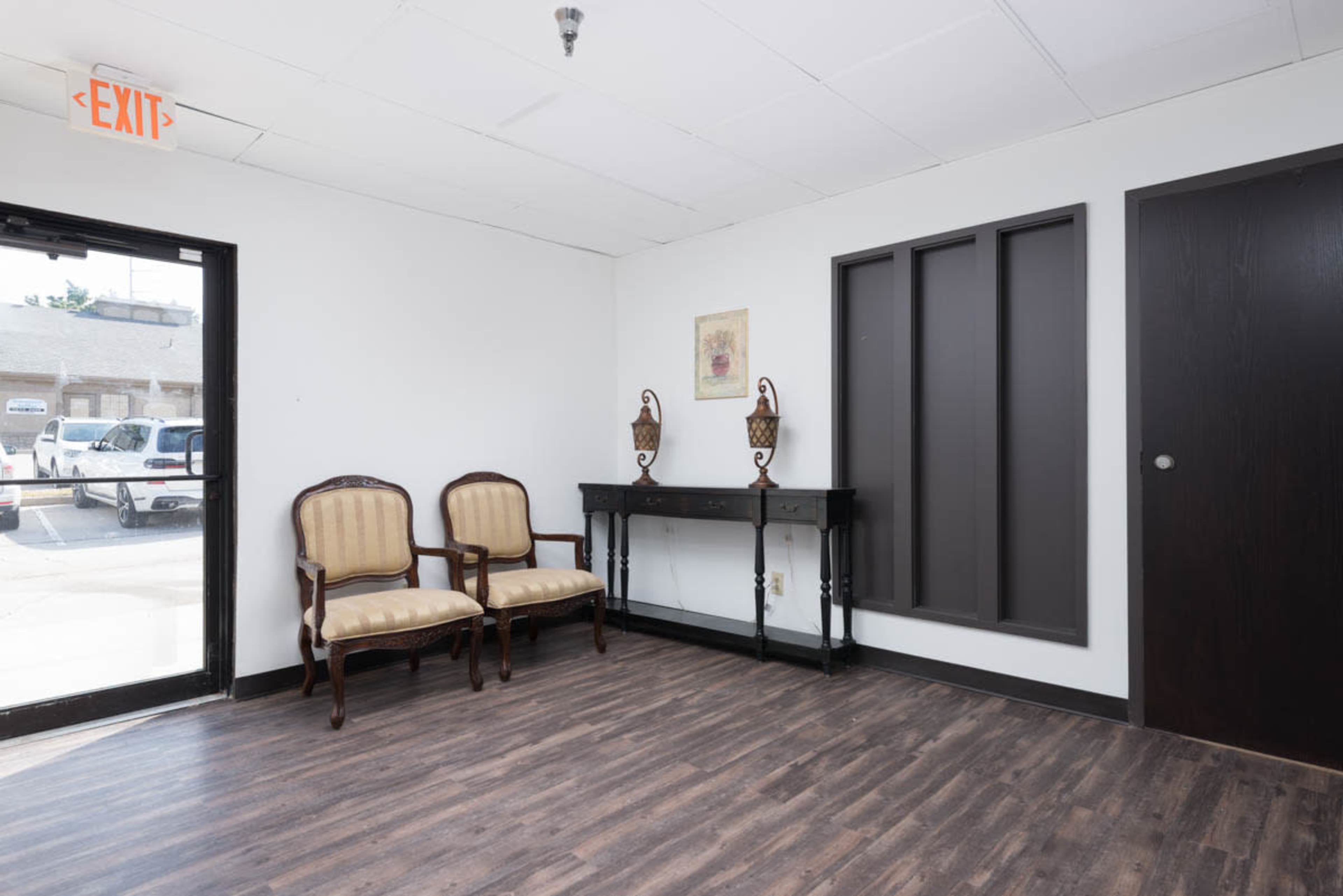 The image shows a sparse foyer with two chairs and a console table against a white wall, featuring an exit sign above the door.