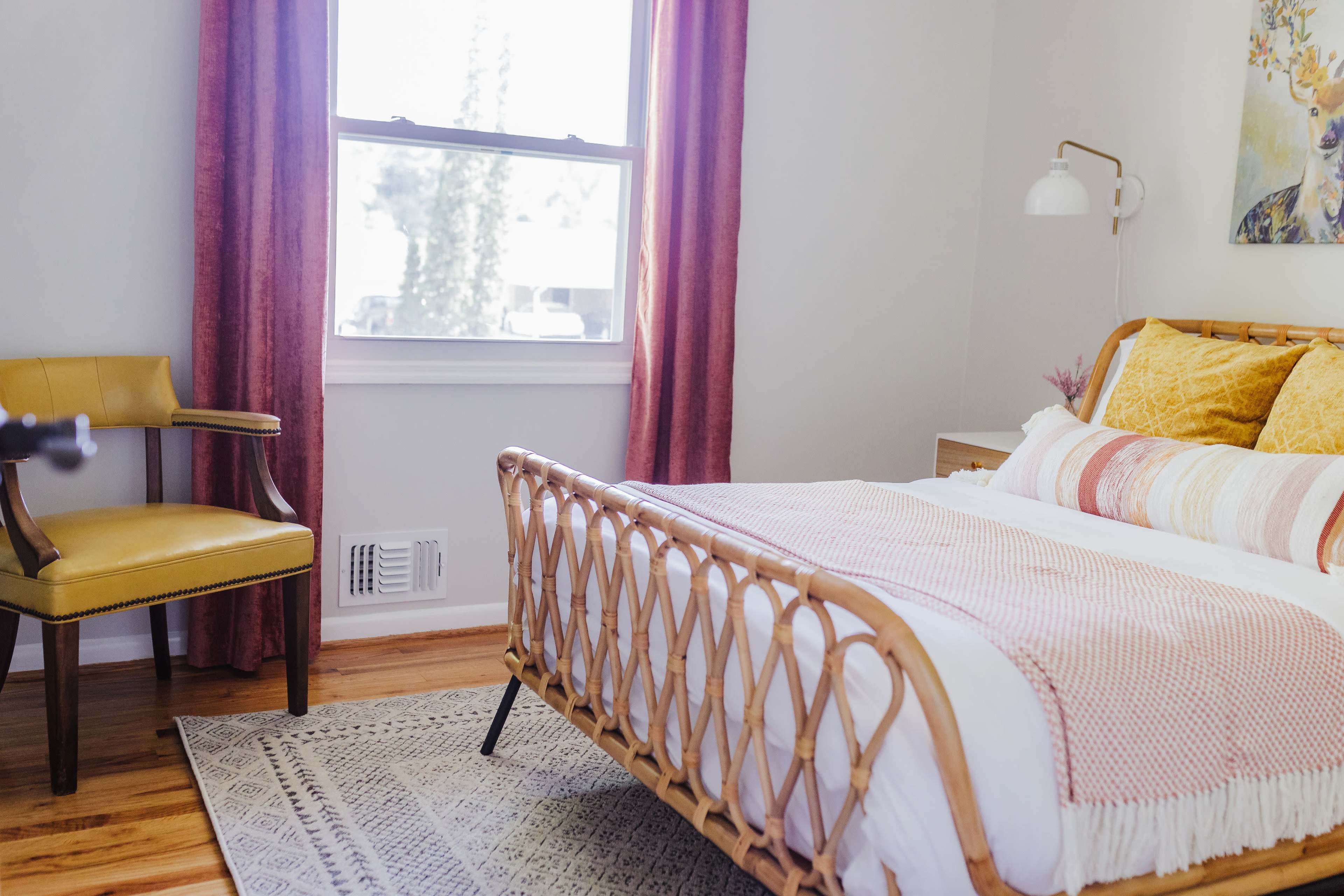 A bedroom with a rattan bed, yellow accent pillows, a wooden chair, and pink curtains, illuminated by natural light from a window.