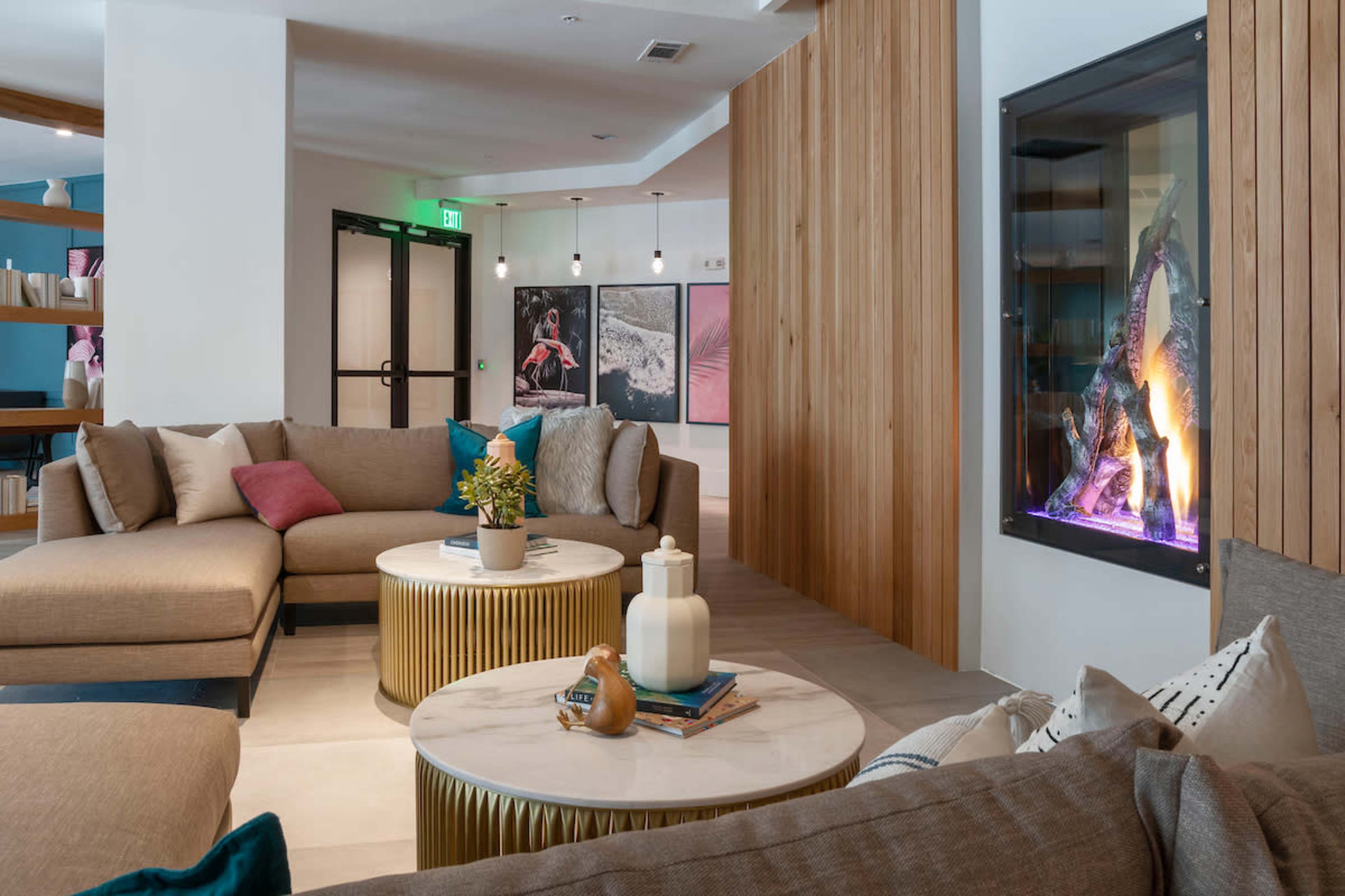 The image shows a modern lounge area featuring a sectional sofa, round coffee tables, and a fireplace embedded in a wooden wall.