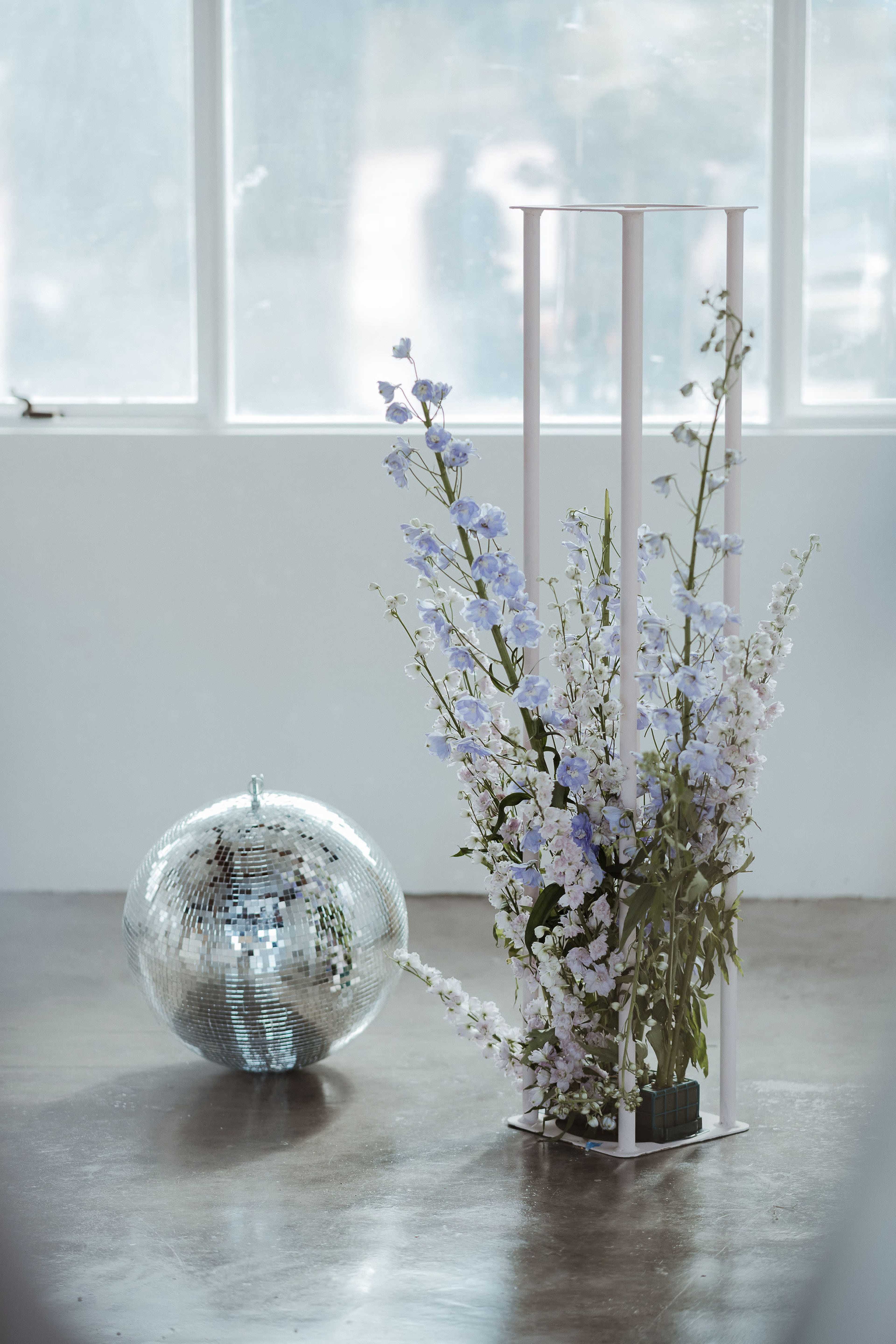 A tall, modern flower arrangement with blue and white flowers stands beside a reflective disco ball in a bright, minimalist room.