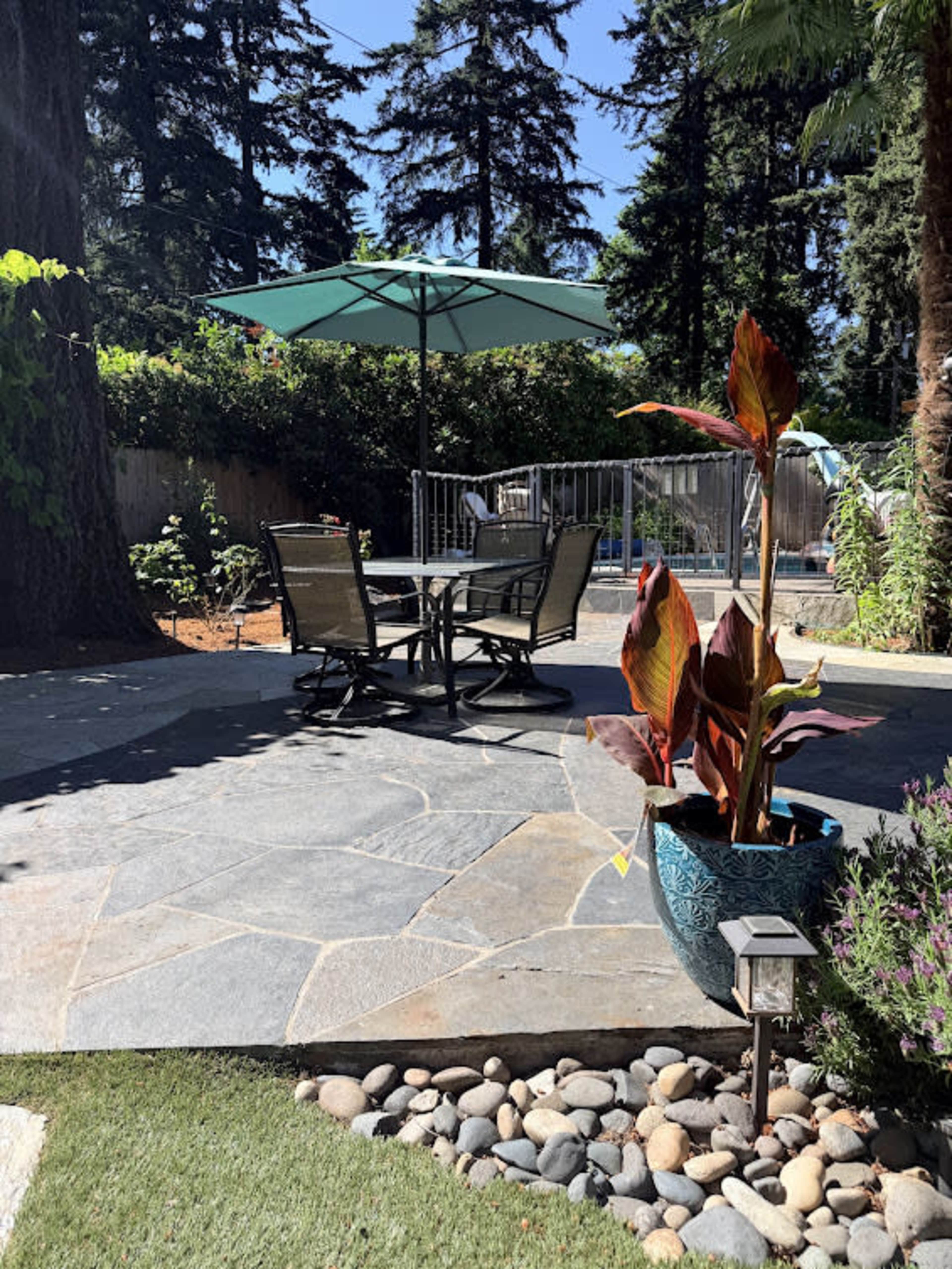 The image shows a patio area with a stone floor, a table and chairs set under a green umbrella, surrounded by plants and trees.
