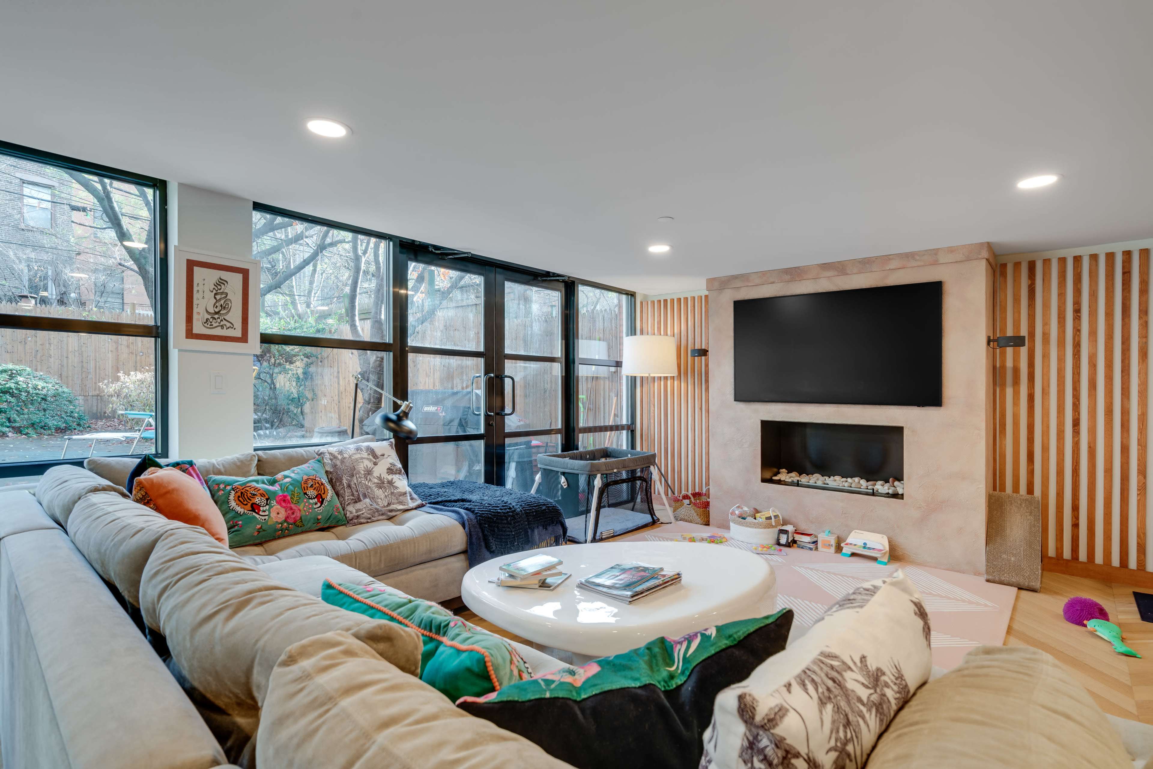A modern living room features a large sectional sofa, a round coffee table, and a mounted television above a fireplace, with large windows providing views of an outdoor space.