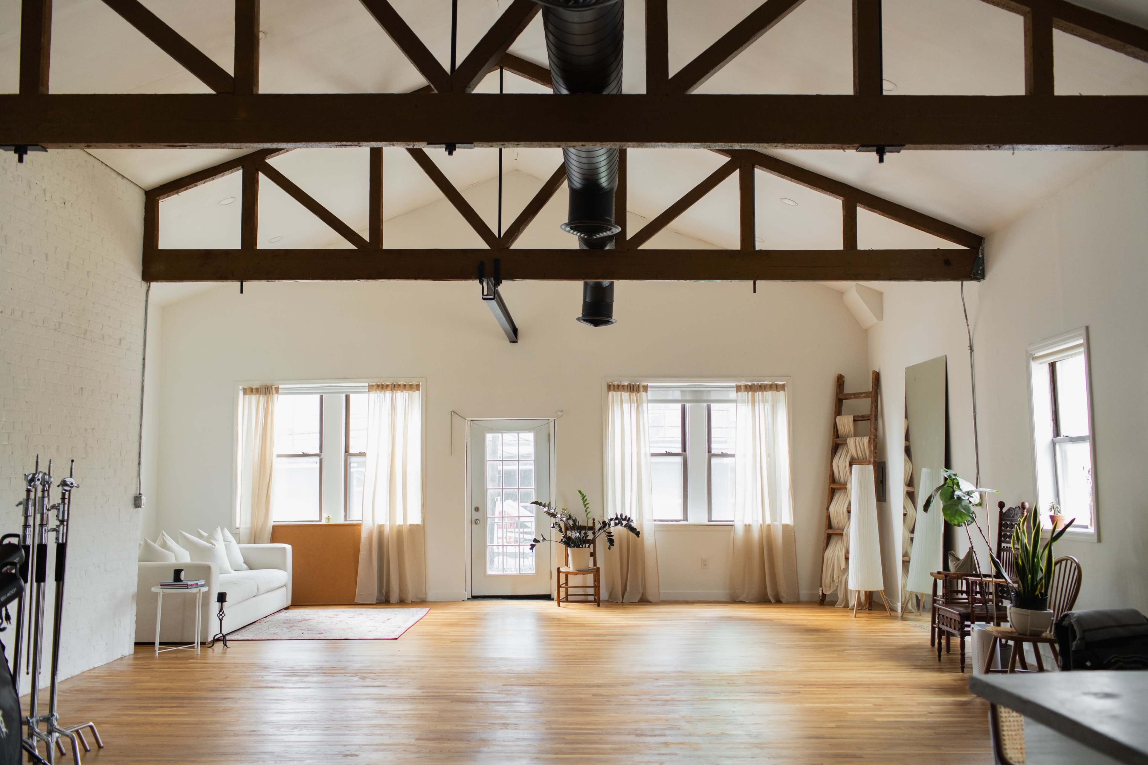 A spacious, well-lit studio with wooden beams, large windows, and simple furnishings including a sofa, chairs, and plants.