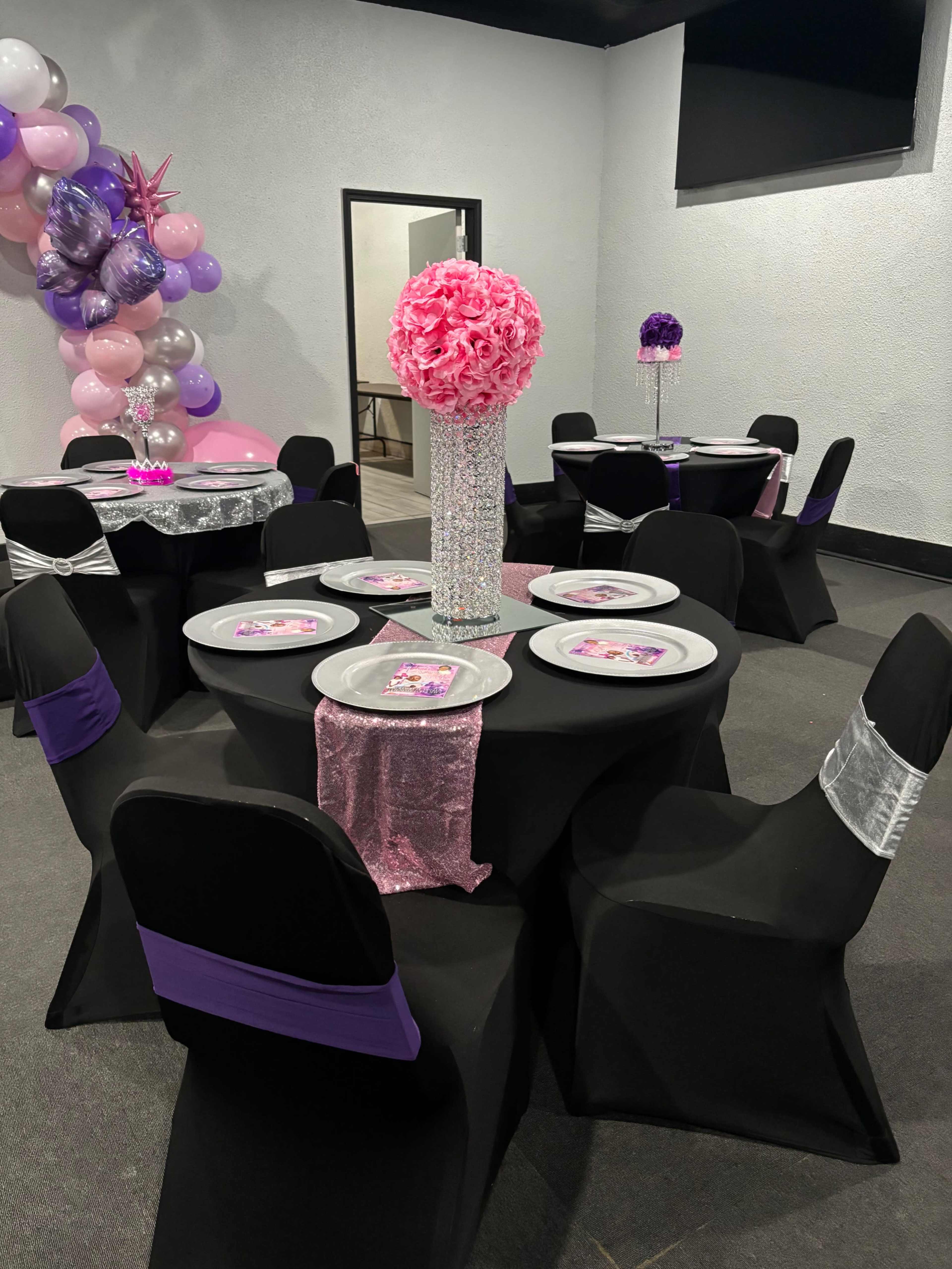 The image shows a decorated event space with black tablecloths, chairs covered in black with purple and silver accents, and floral centerpieces.