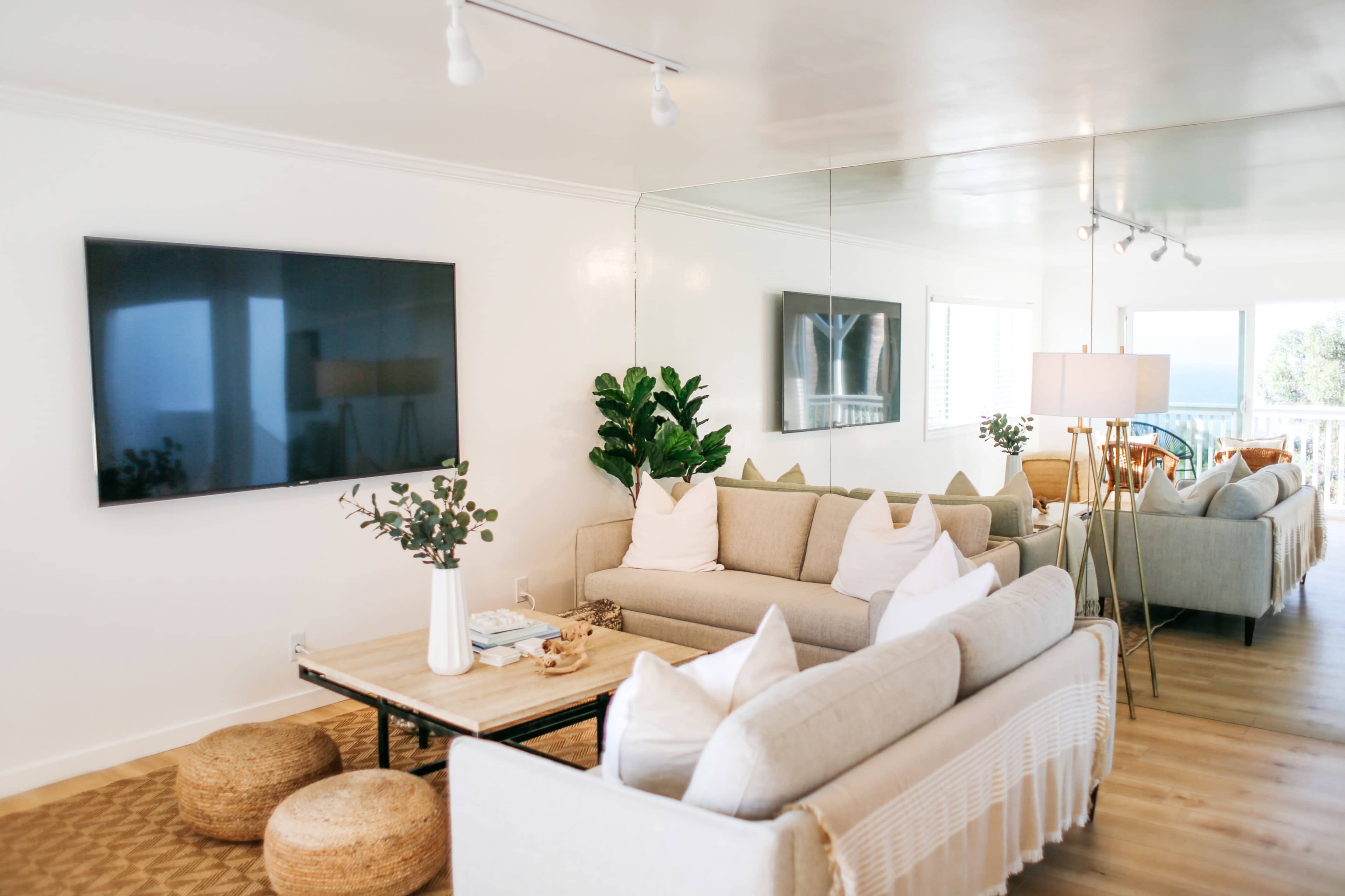 A modern living room with a light color scheme, featuring two sofas, a coffee table, and a large wall-mounted TV, along with plants and decorative elements.