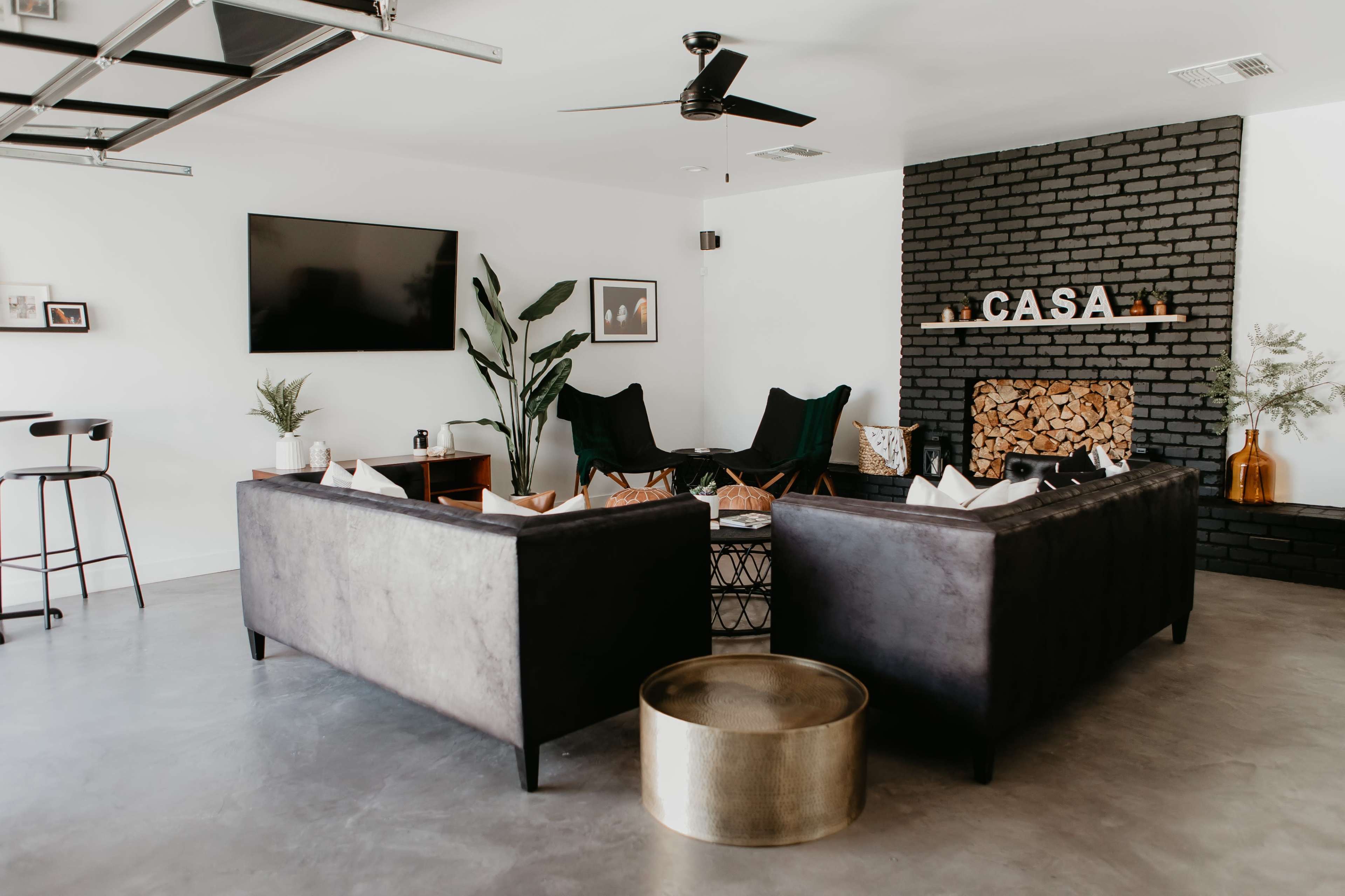 A modern living room features two dark couches facing each other, a coffee table in the center, a wall-mounted TV, and a decorative fireplace with the word "CASA" above it.