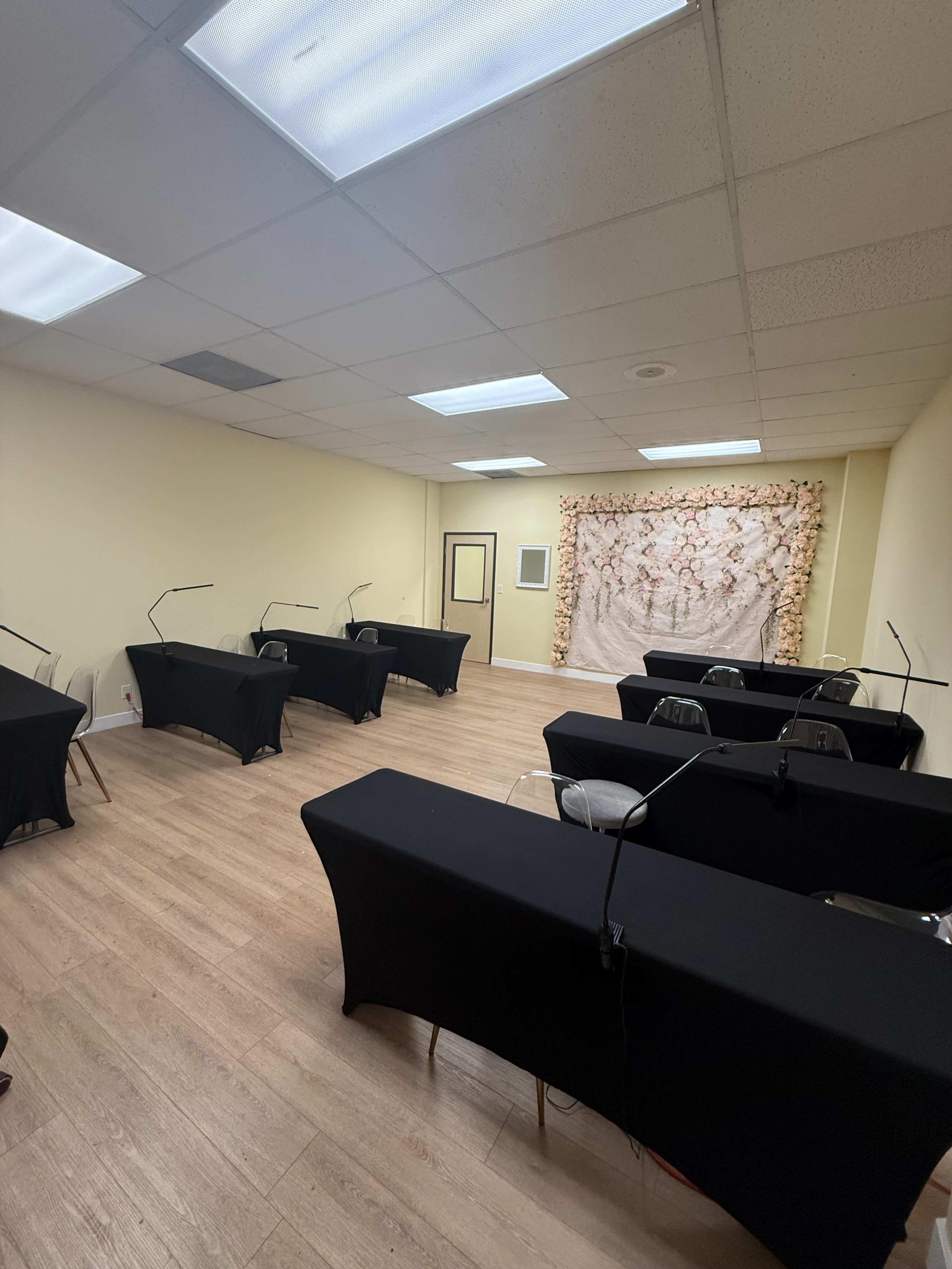 The image shows a spacious room set up with several tables covered in black cloth, each equipped with a reading lamp, and a backdrop adorned with floral decorations.