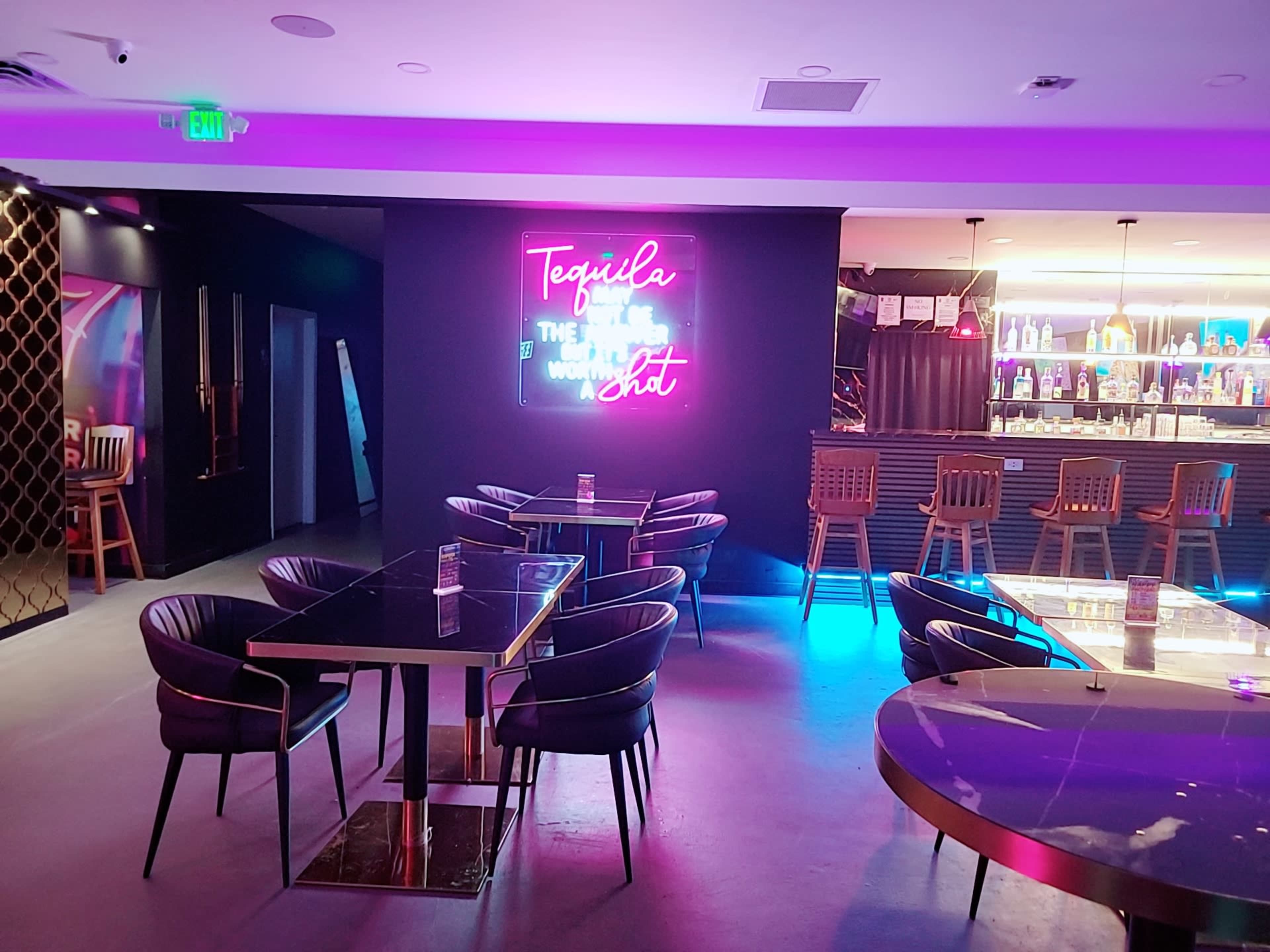 The image shows a modern bar area with black chairs, illuminated tables, and a neon sign on the wall that reads "Tequila" along with a brightly lit bar in the background.