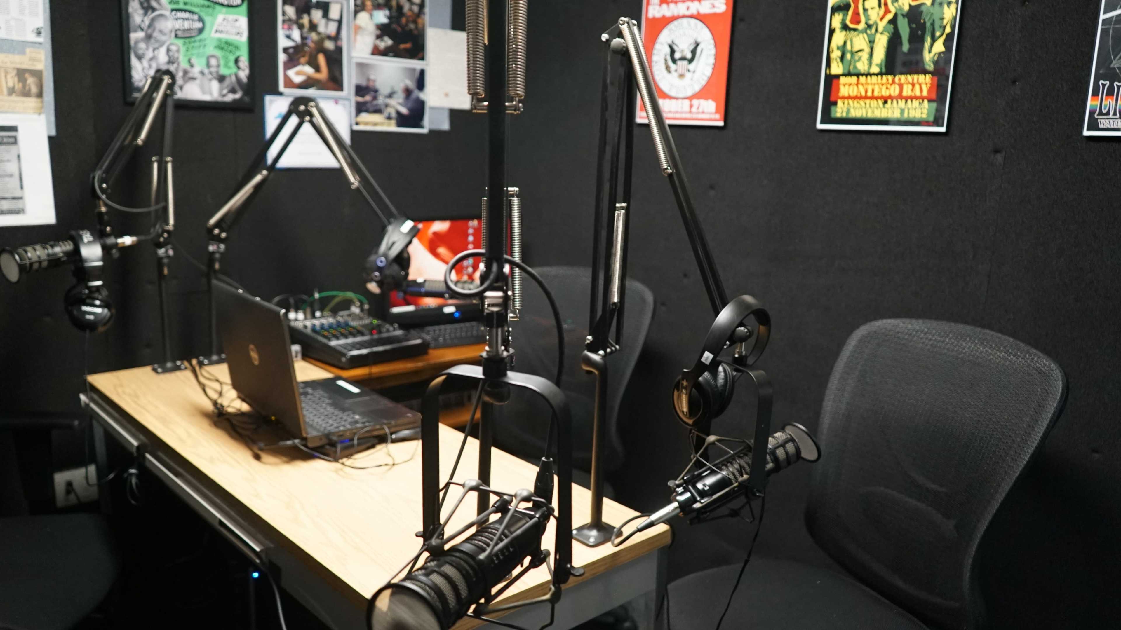 The image shows a radio studio setup with several microphones, a laptop, audio equipment, and chairs in a dimly lit, soundproofed space.