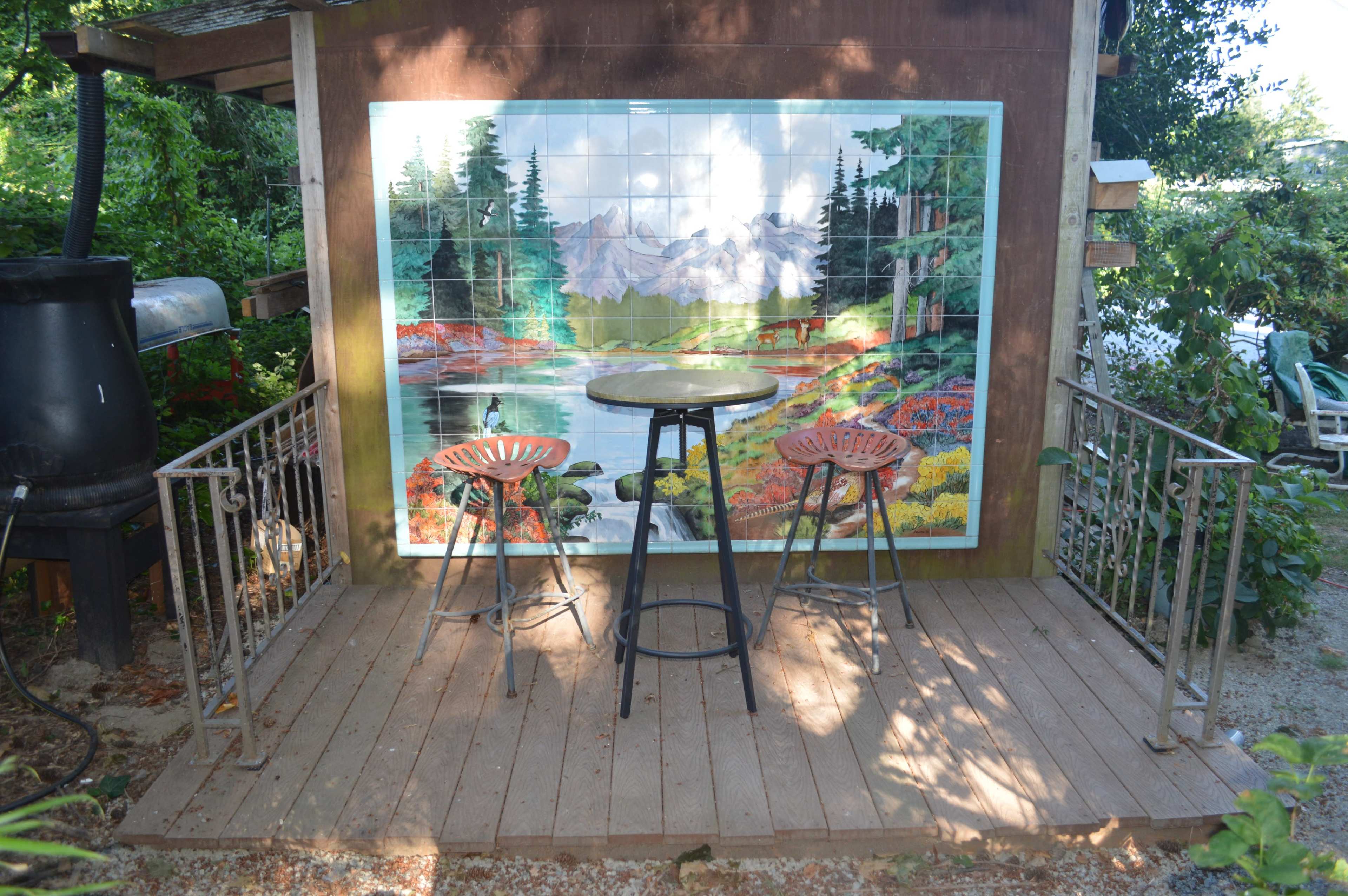 A wooden deck with two stools faces a mural of a natural landscape, surrounded by greenery.