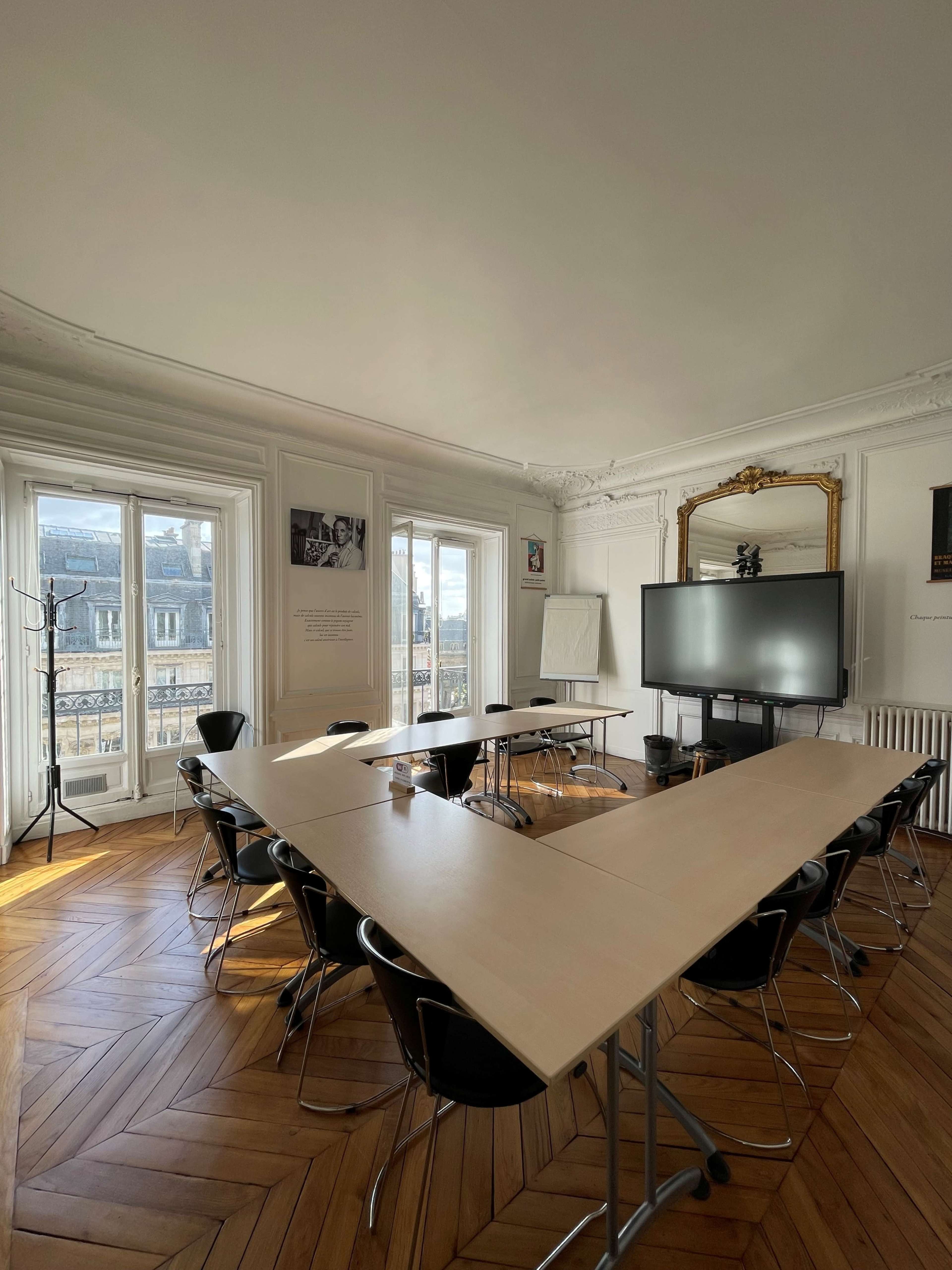 The image shows a conference room with a large table arranged in a triangular formation, black chairs surrounding it, and a screen on one side, illuminated by natural light from large windows.
